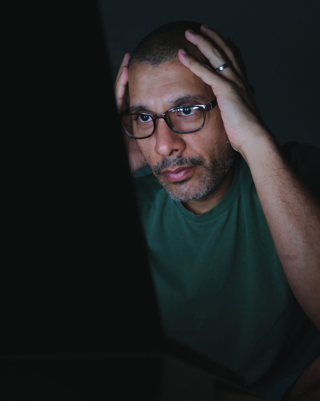 Man with glasses holding his head, looking at a laptop in a dimly lit room.
