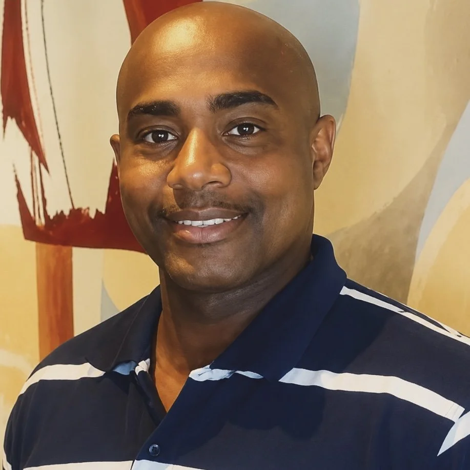 Close-up of a smiling man with a shaved head wearing a navy blue and white striped polo shirt, standing in front of an abstract colorful wall.