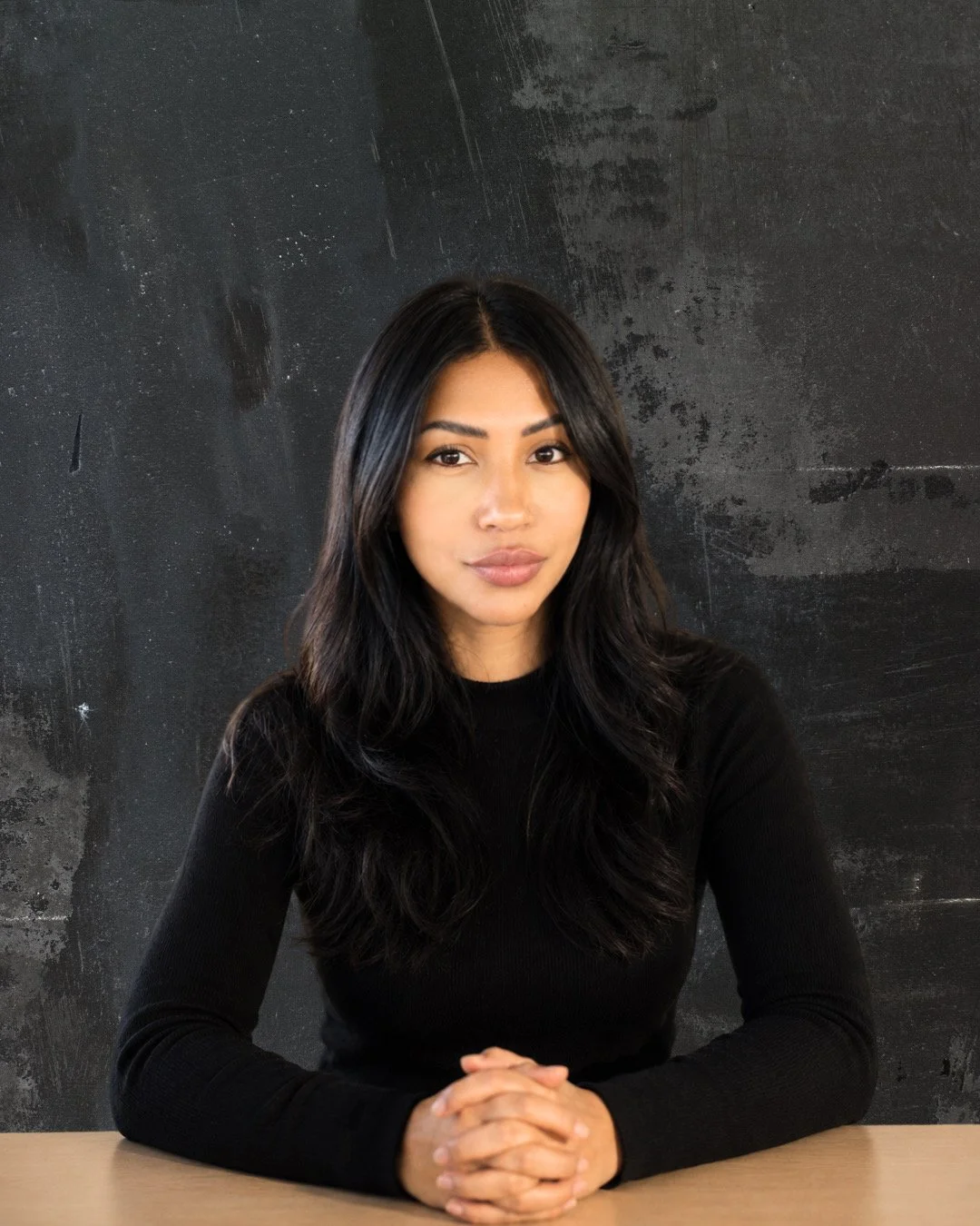 Tassja Cadoch with long black hair, wearing a black top, sitting at a table with hands clasped, in front of a dark textured wall.
