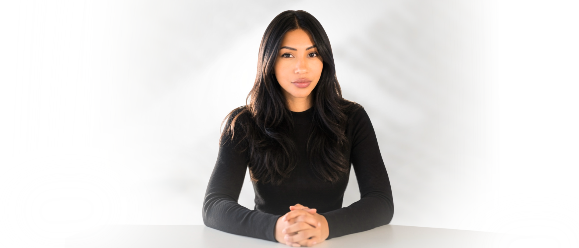 A woman with long black hair, wearing a black long-sleeve top, sitting at a white table against a white background with a subtle shadow behind her.