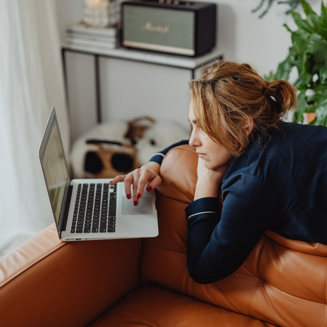 A woman with red hair lying on her stomach on a brown leather couch, looking at her laptop screen with a focused expression. A dog is resting in the background.