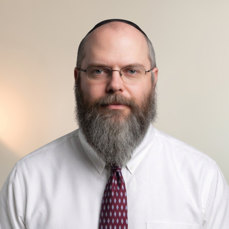 Rabbi Alan Friedlander with glasses, a full beard, wearing a white shirt and a purple patterned tie, looking at the camera against a plain, light-colored background.