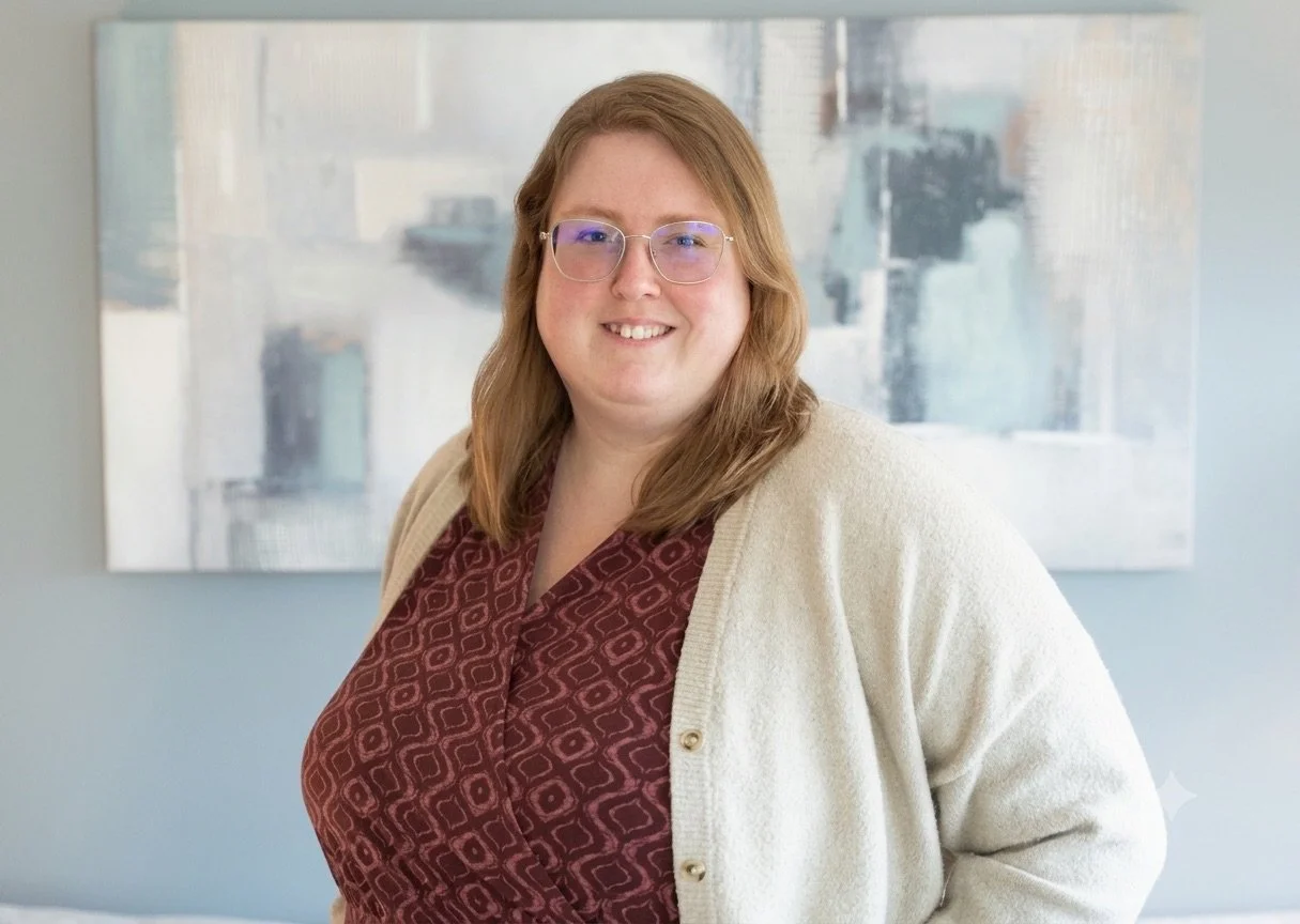 A woman with shoulder-length light brown hair, glasses, and a beige cardigan over a patterned maroon blouse, smiling in front of an abstract painting with soft pastel colors.