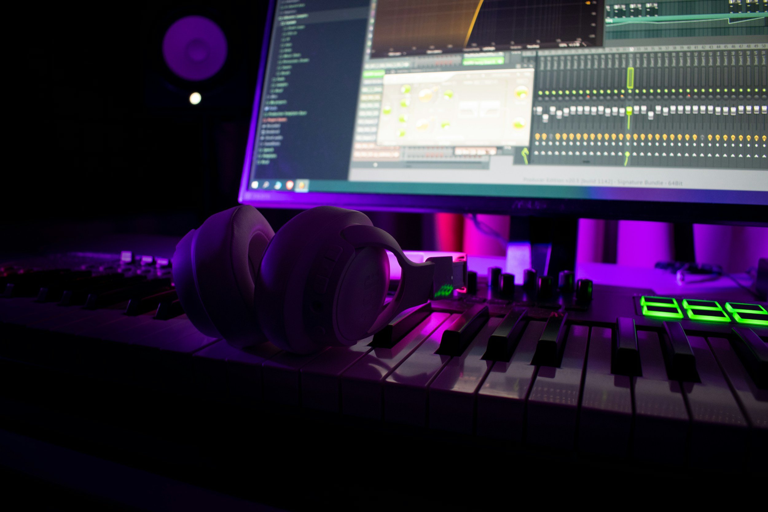 A computer monitor displaying digital audio workstation software with colorful audio tracks and controls. In front of the monitor is a keyboard, and a pair of white headphones rests on the keyboard. The scene is illuminated with purple and pink lighting, creating a vibrant workspace.