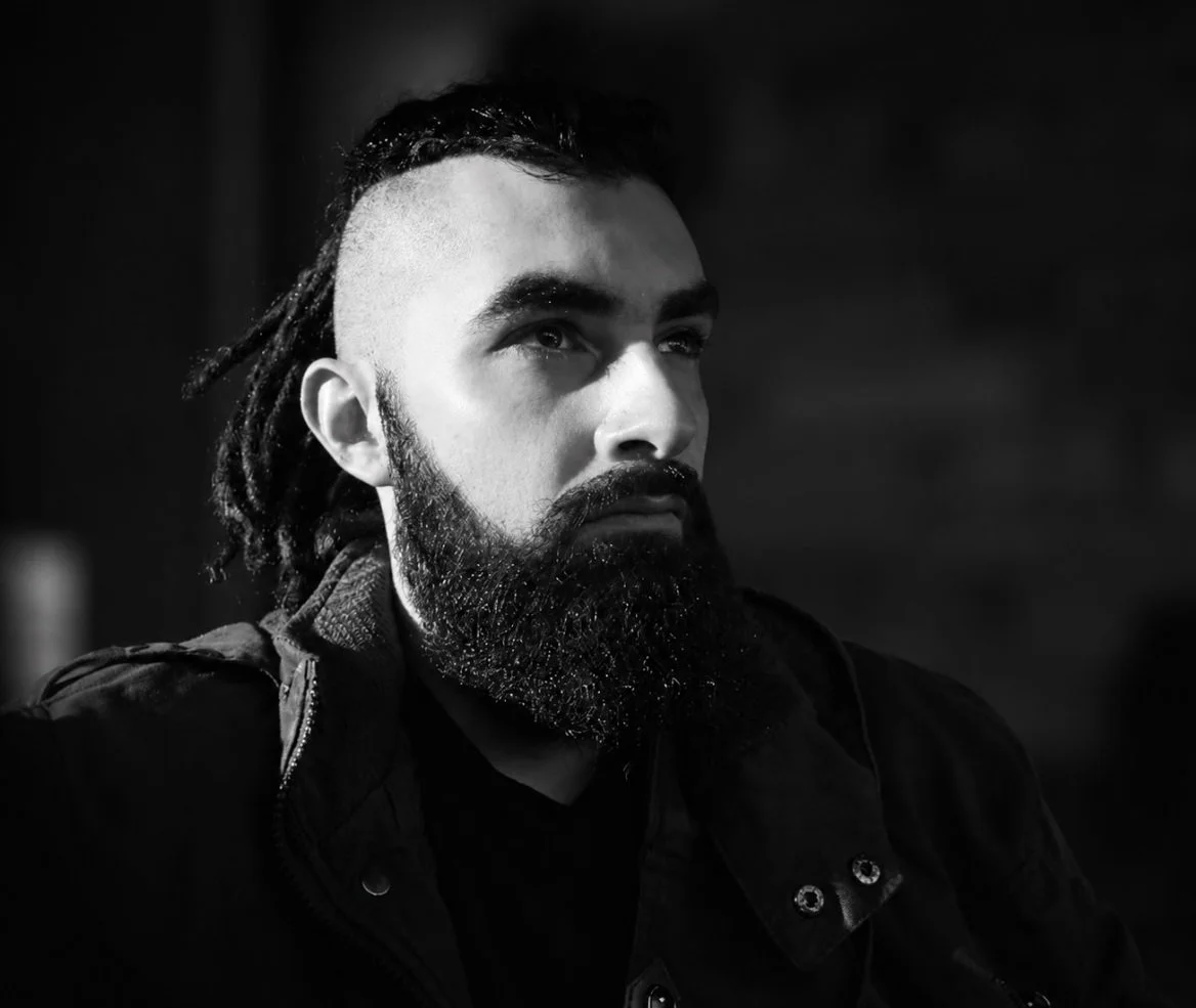 A black and white photo of a man with a beard and dreadlocks, wearing a dark jacket, looking upwards with a serious expression.