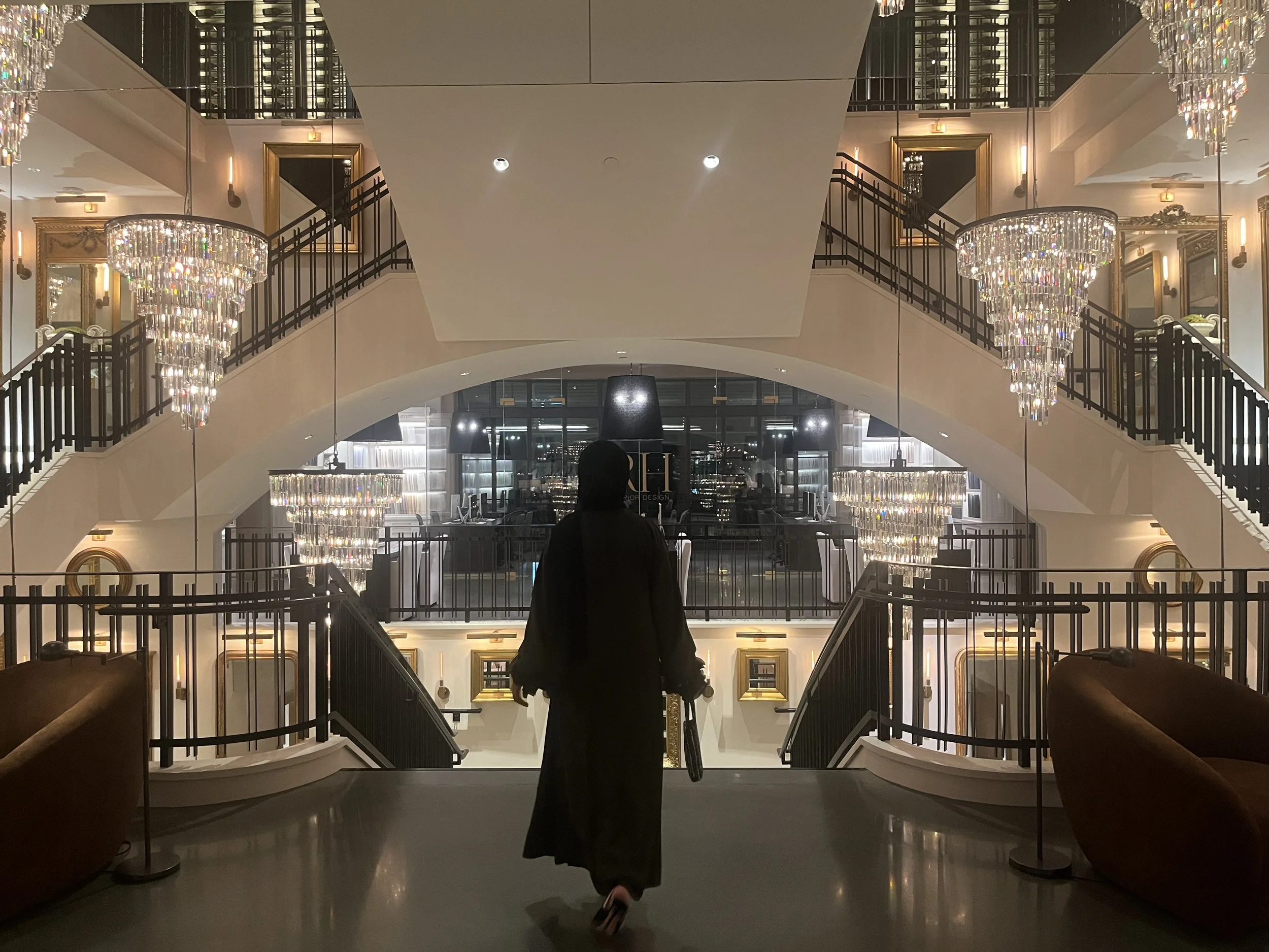 A person with long hair, wearing a black dress, standing at the bottom of a grand staircase inside a luxurious building. The staircase has black metal railings and is illuminated by large, elegant chandeliers hanging from the ceiling. The interior features gold-framed mirrors and art on the walls, with modern lighting fixtures.