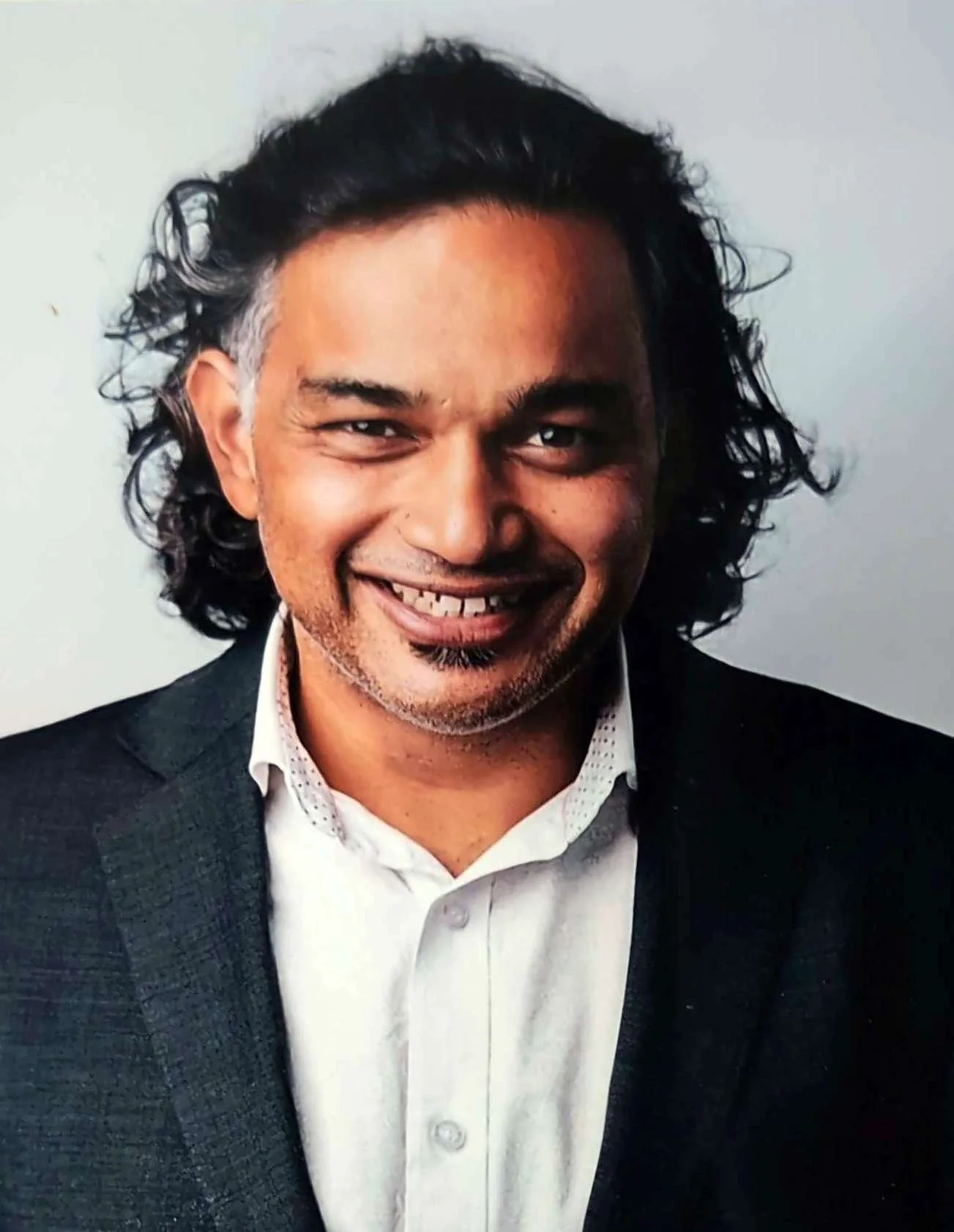 A smiling man with curly black hair, wearing a dark suit jacket over a white collared shirt, against a plain light background.