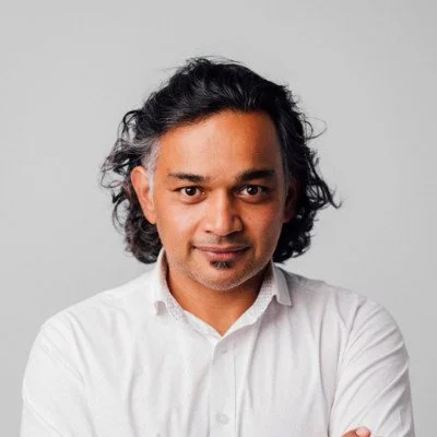 A man with curly dark hair and a slight smile, wearing a white button-up shirt, looking at the camera against a plain light gray background.