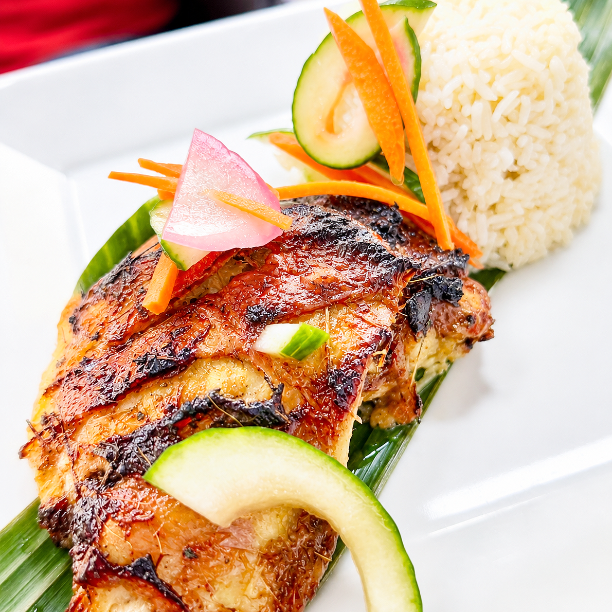 Roasted Bacolod-style Chicken Inasal topped with sliced radish, cucumber, and carrot, served with a side of rice and garnished with lemon and green onion; plated on a white dish with banana leaf decorations.