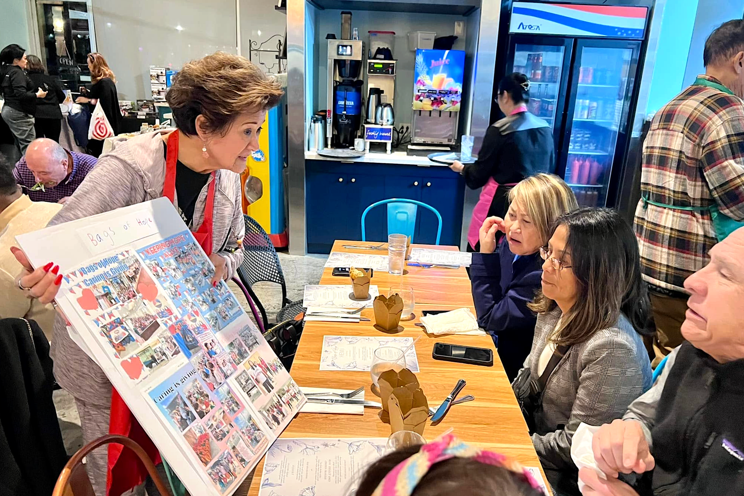 People gathered around a table at a restaurant or cafe, with a woman in the foreground holding a colorful collage or poster. The table has menus, glasses of water, and food items, and a large digital display and beverage fridge are visible in the background.