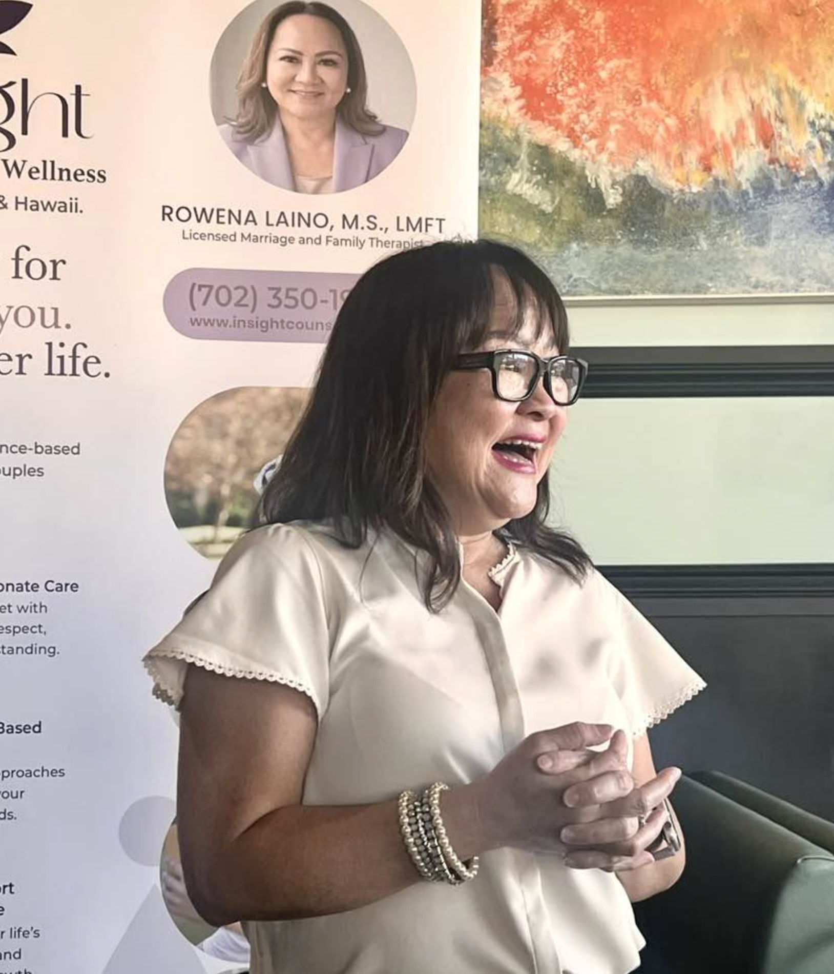 A woman with dark hair and glasses, smiling while holding a phone, standing in front of a poster with her photo and contact information, and a colorful abstract painting in the background.
