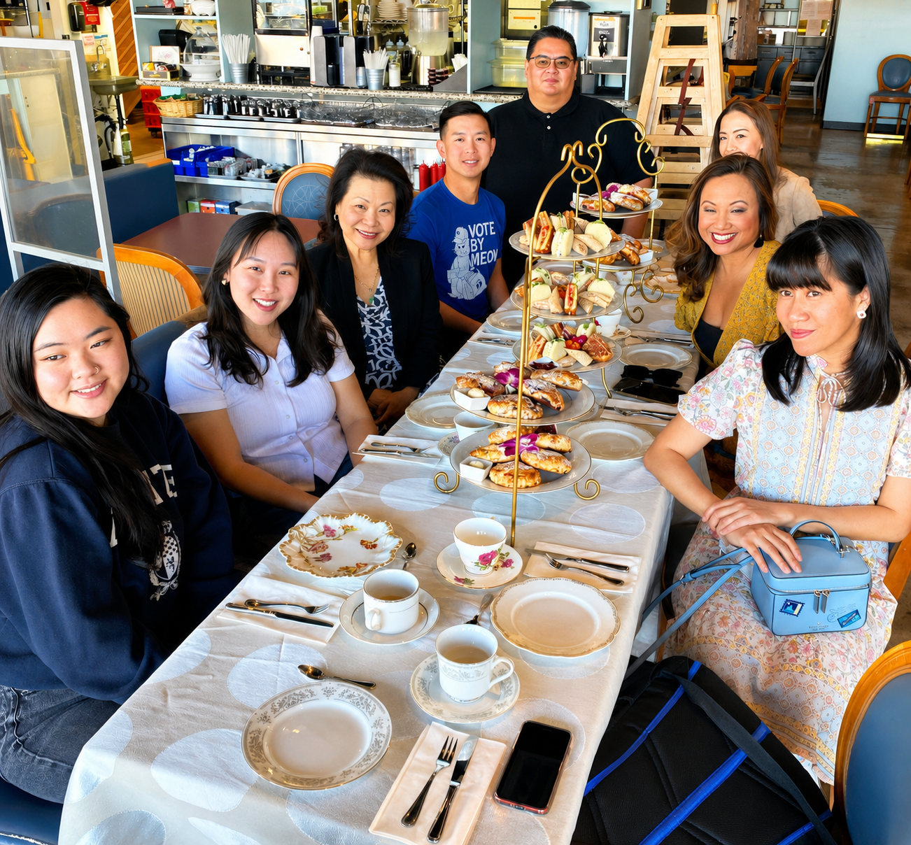 A group of eight women and two men sitting at a table set for afternoon tea, with tiered trays of sandwiches, pastries, and desserts in a restaurant or cafe.