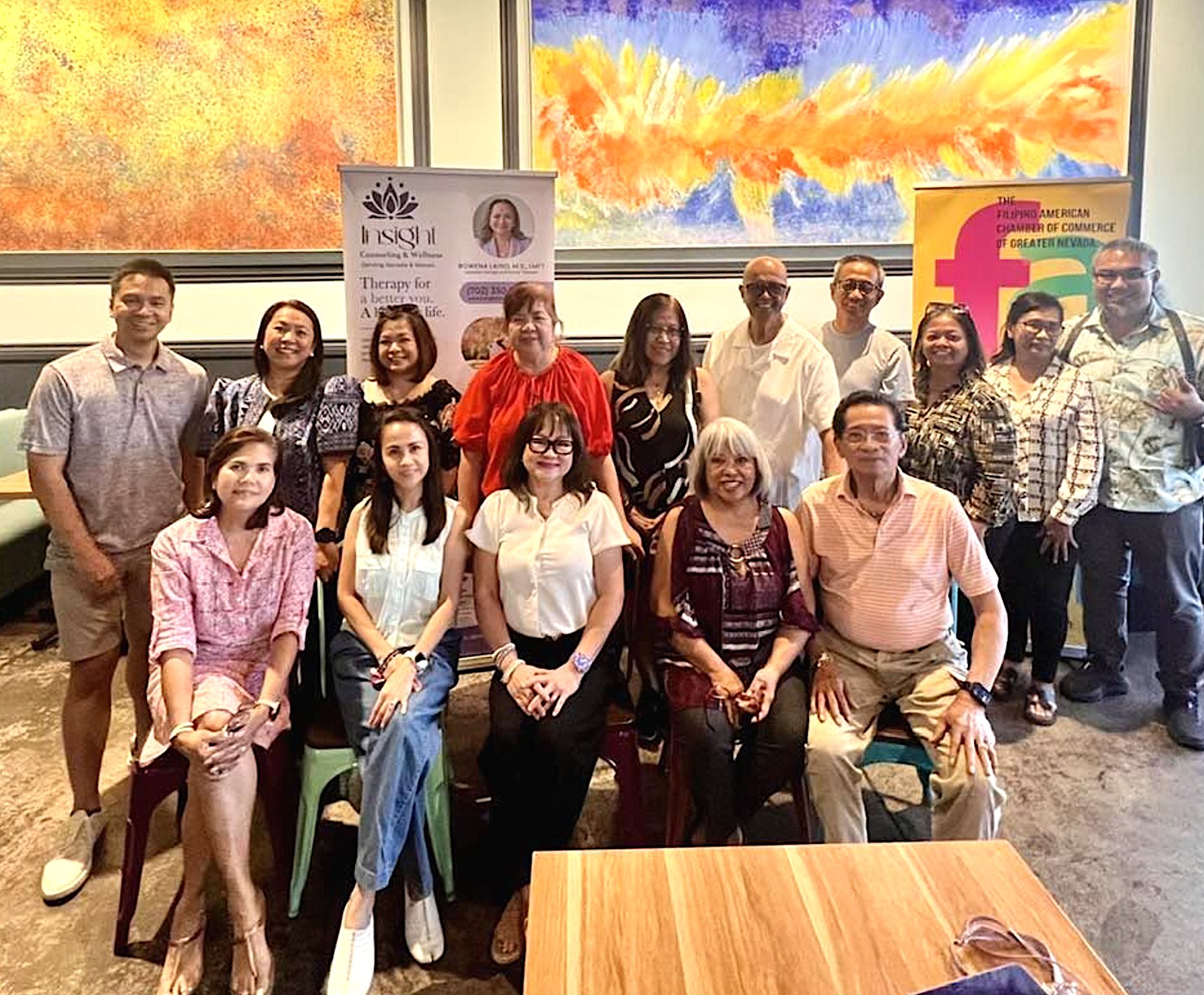 A group of people gathered together in a room, posing for a photo. Behind them are colorful abstract paintings and banners related to a Filipino-American chamber of commerce event.