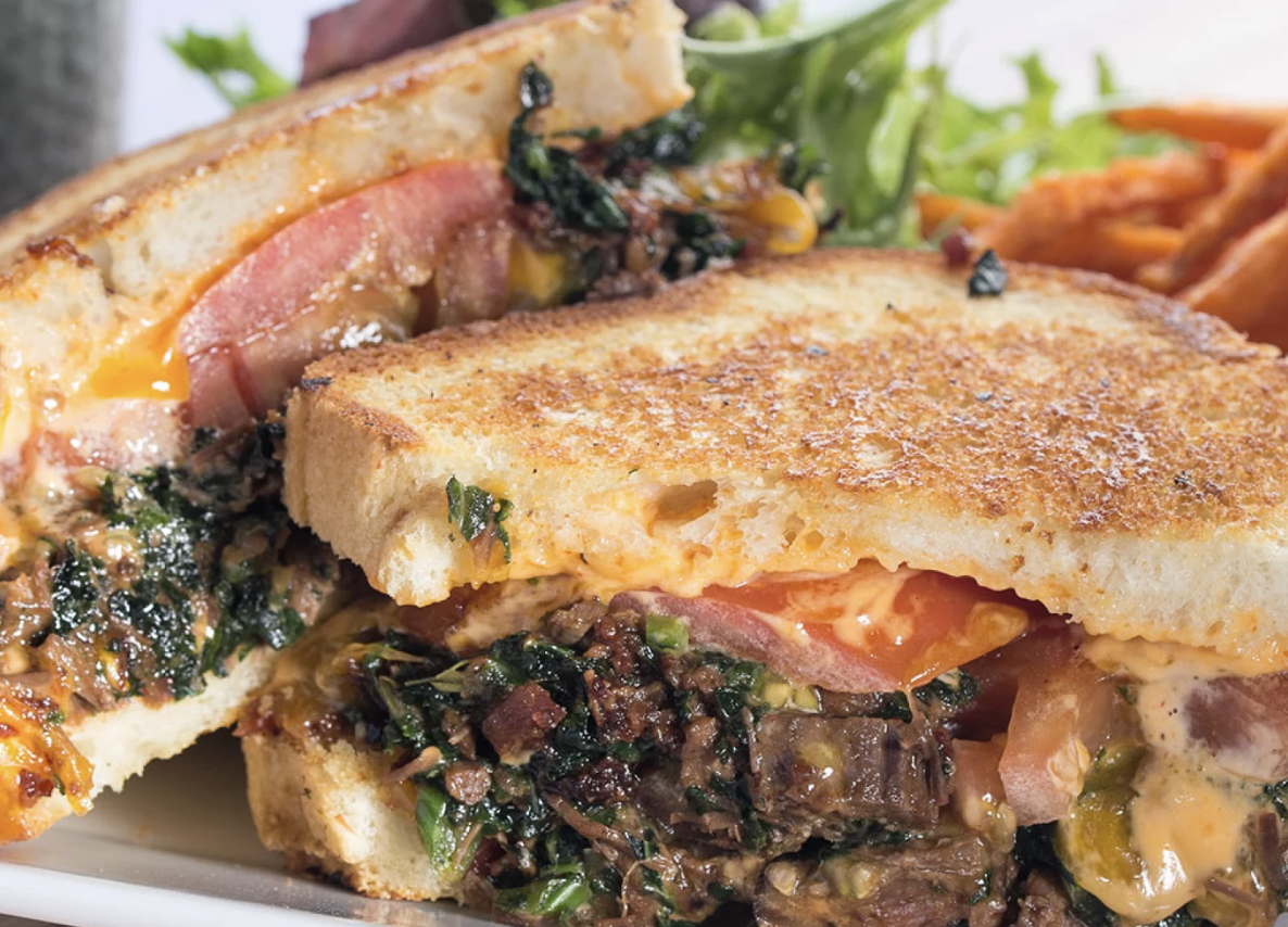 Close-up of a toasted sandwich with melted cheese, ham, tomato, greens, and cooked vegetables inside, served with a side salad.