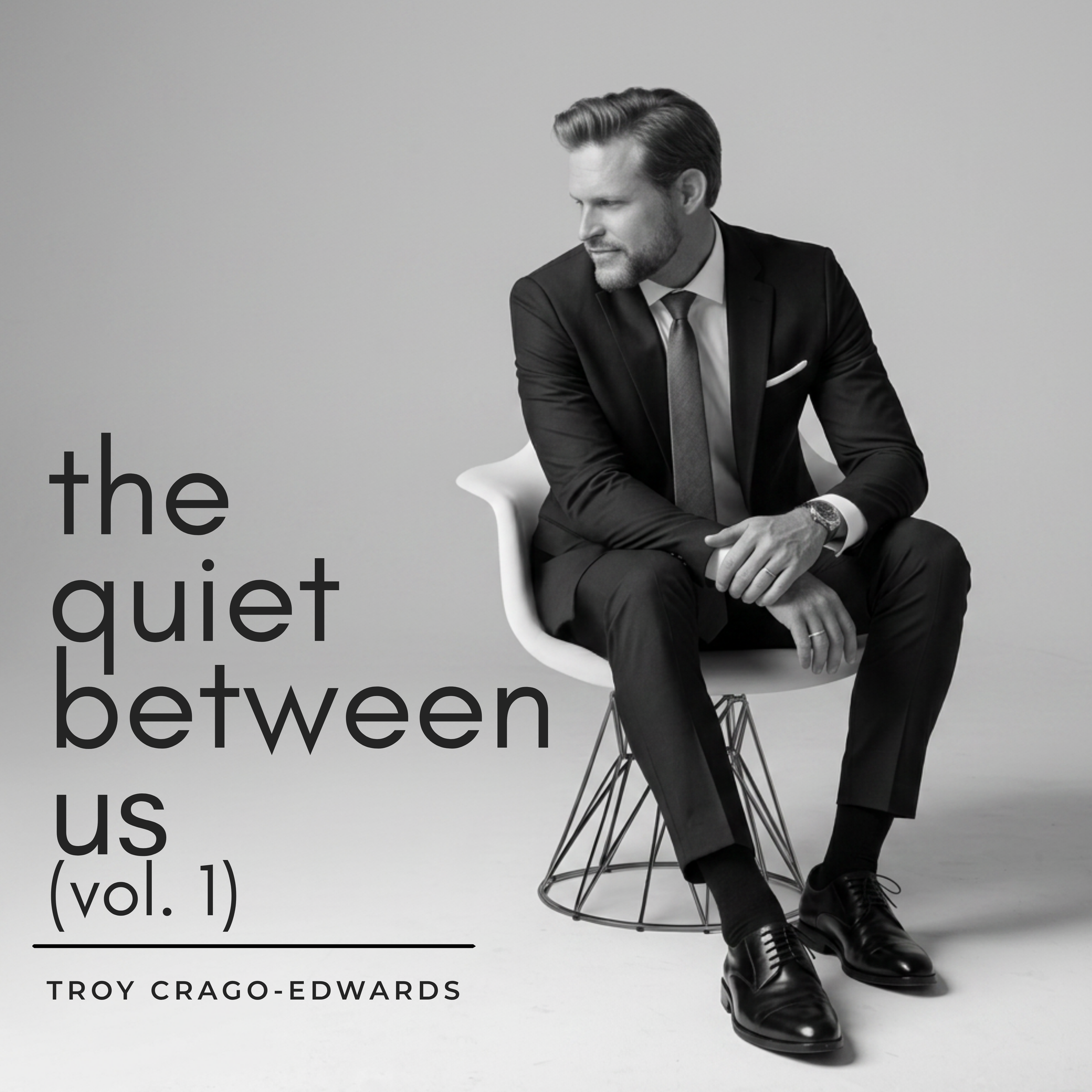 Black and white photograph of a man in a suit sitting on a modern chair against a plain background, with large text overlaying part of the image reading 'the quiet between us (vol. 1)' and 'TROY CRAGO-EDWARDS' beneath.