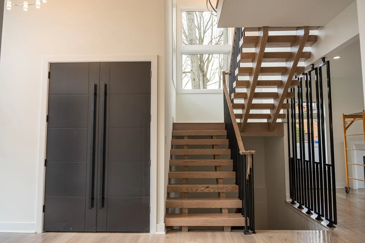 Interior of a modern home with a black double-door entrance on the left, a staircase with wooden steps and black railing in the center, a window above the staircase, and a yellow scaffold on the right.