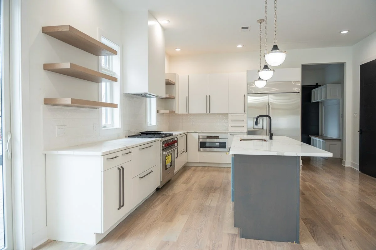 Modern kitchen with white cabinets, stainless steel appliances, wooden floating shelves, marble countertop island, and hardwood flooring.