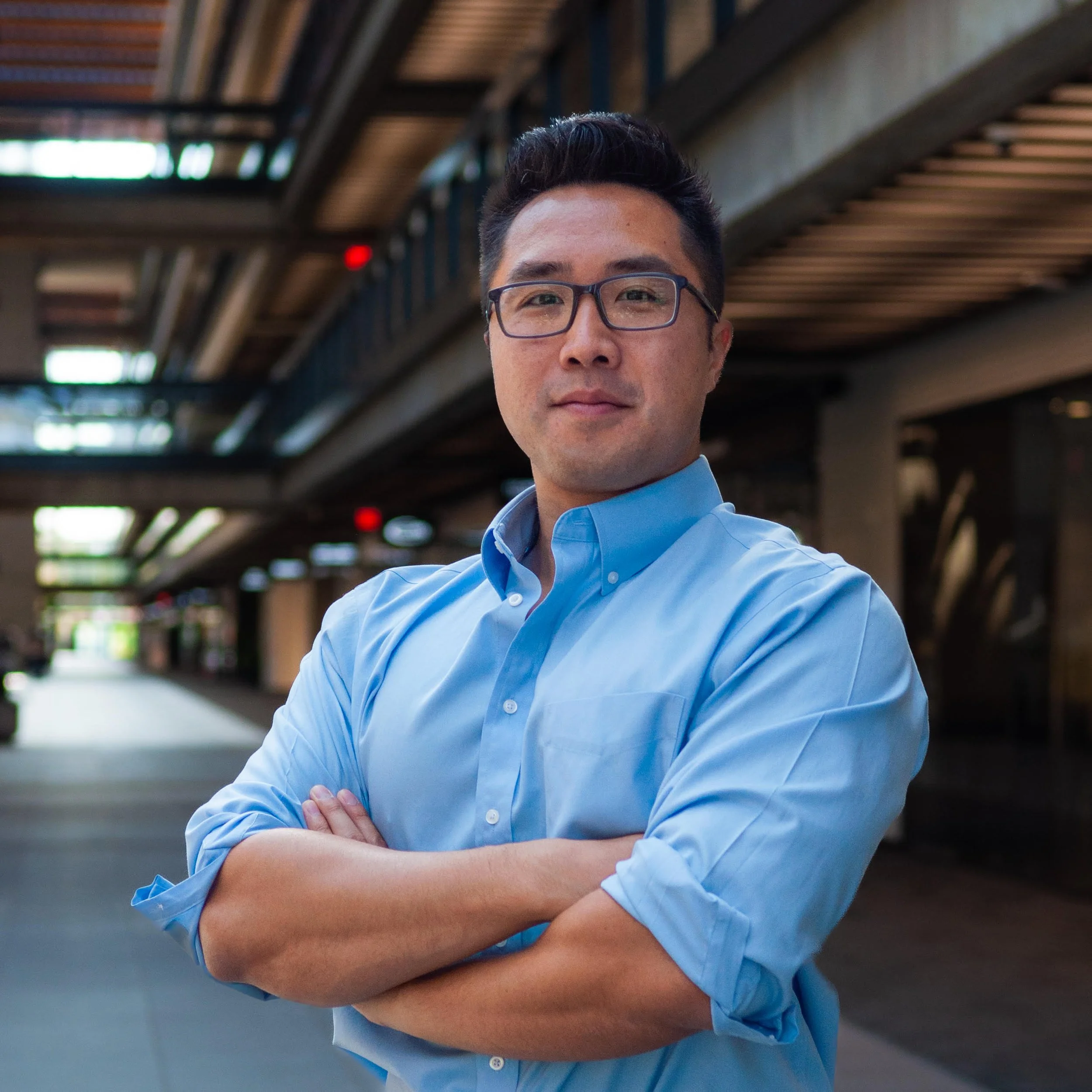 A man with glasses wearing a blue shirt crossing his arms in an urban setting with architectural structures overhead.