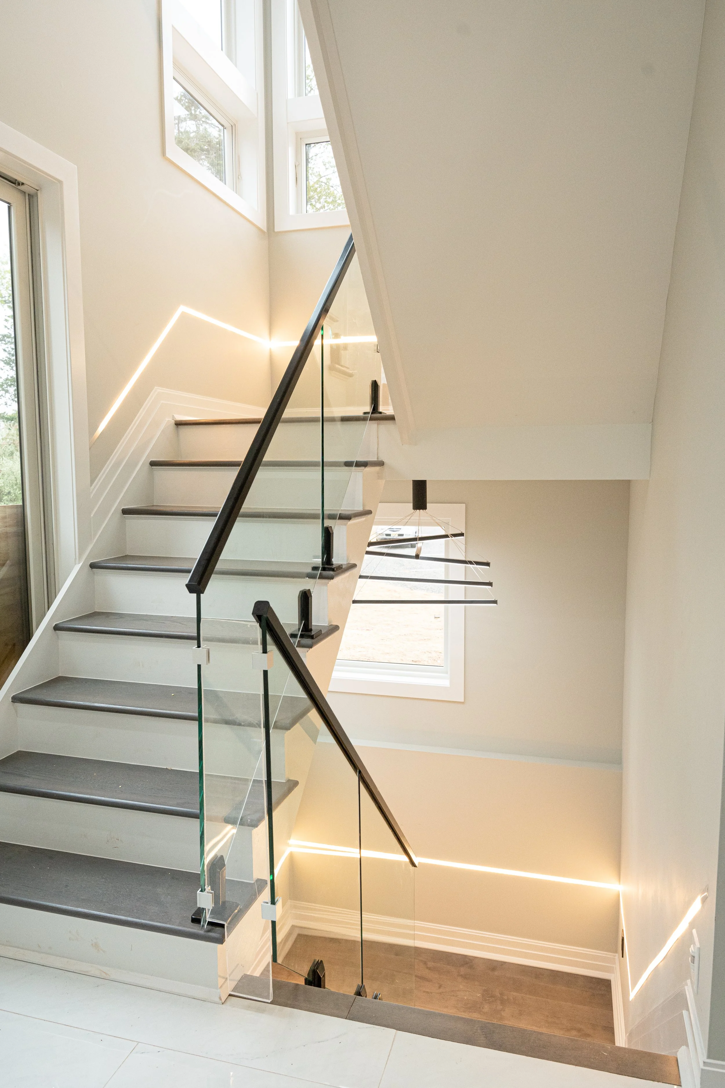 Interior view of a modern staircase with gray steps, glass railing, and large windows letting in natural light, with contemporary lighting accents on the walls.