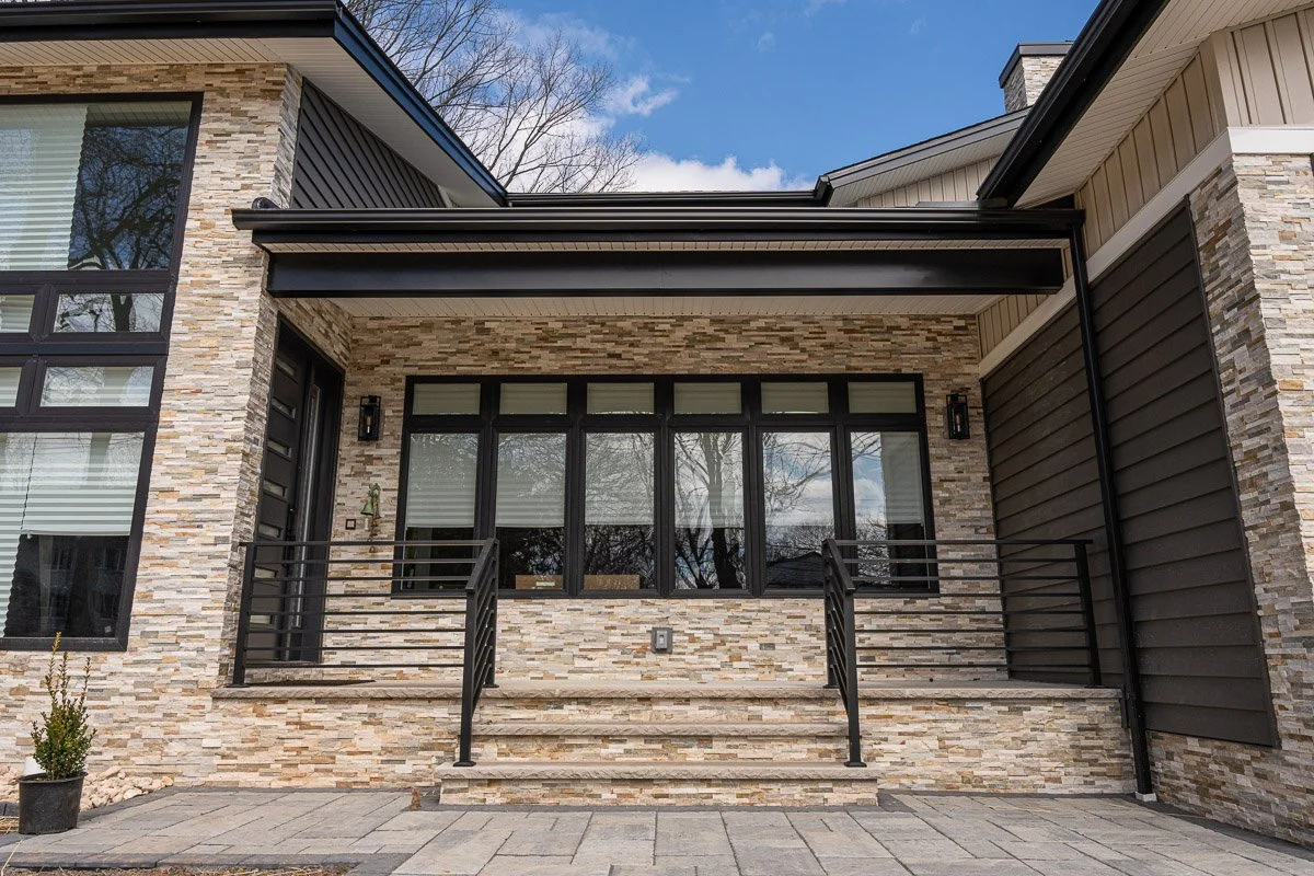 Front entry of a modern house with a stone facade, large black-framed windows, a small balcony with metal railings, and stairs leading up to the door, under a partly cloudy blue sky.