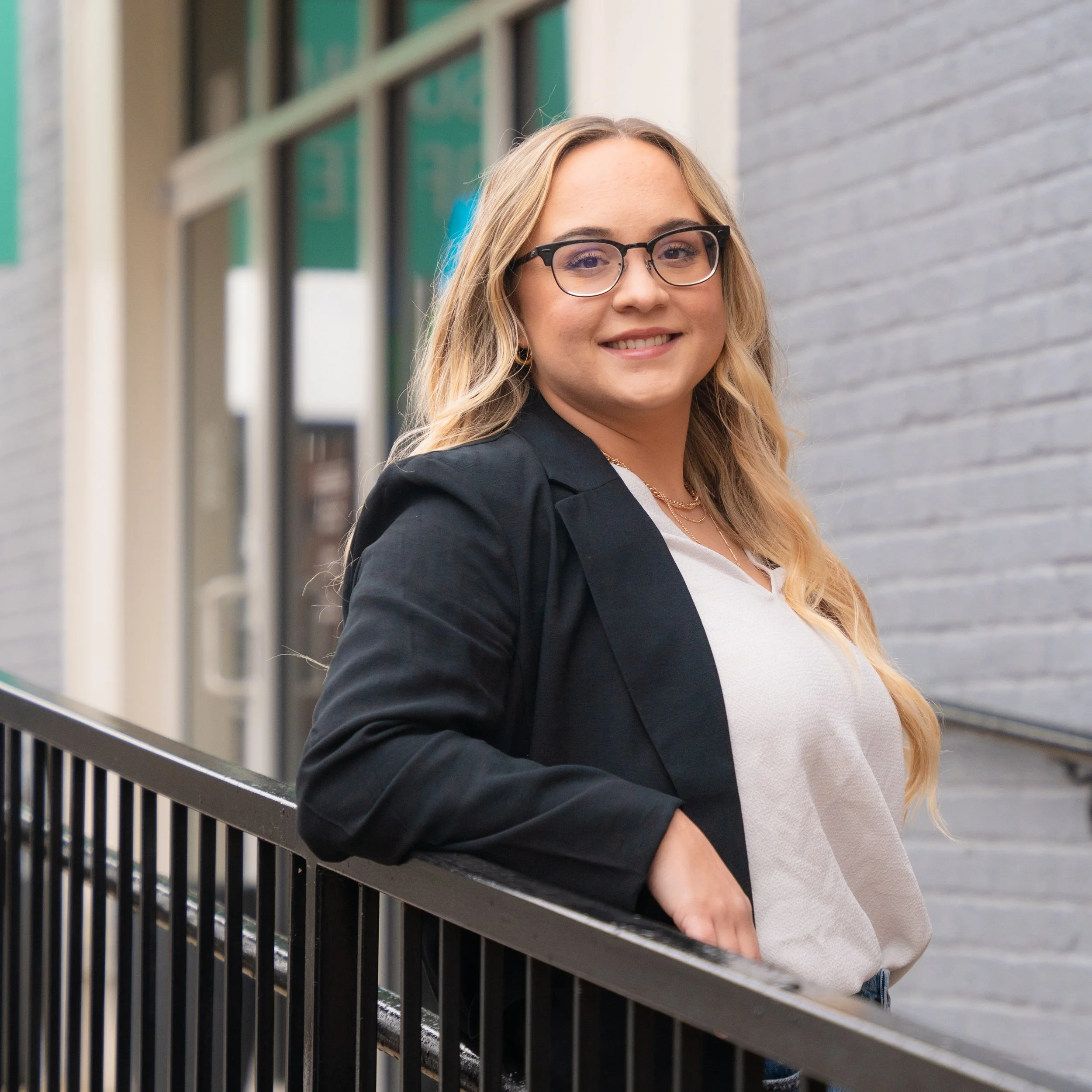 A young woman with blonde wavy hair and glasses smiling and leaning against a metal railing outside near a gray brick wall.