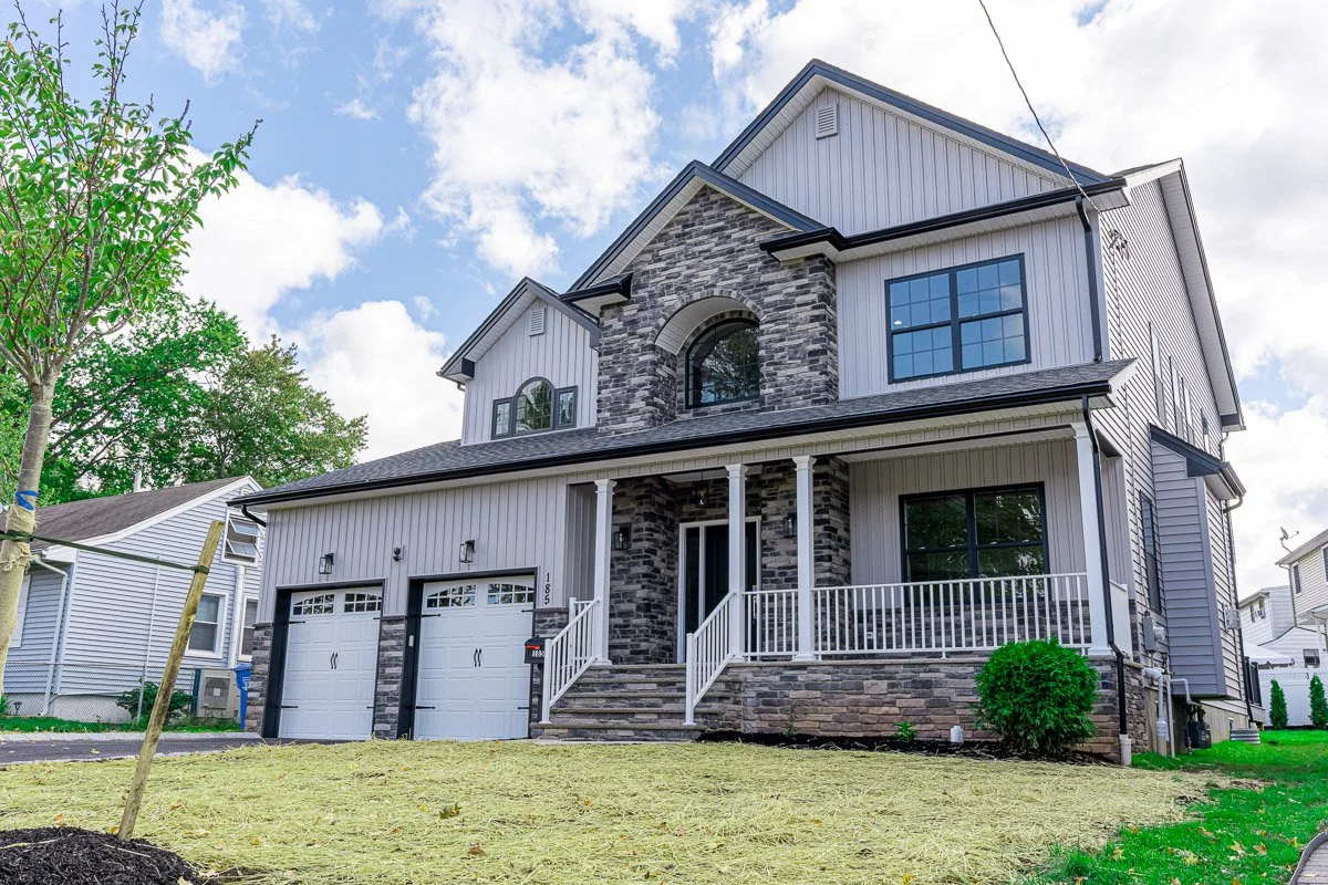 A modern, two-story house with a stone and white siding exterior, front porch with white railing, garage doors, and large windows, under a partly cloudy sky.
