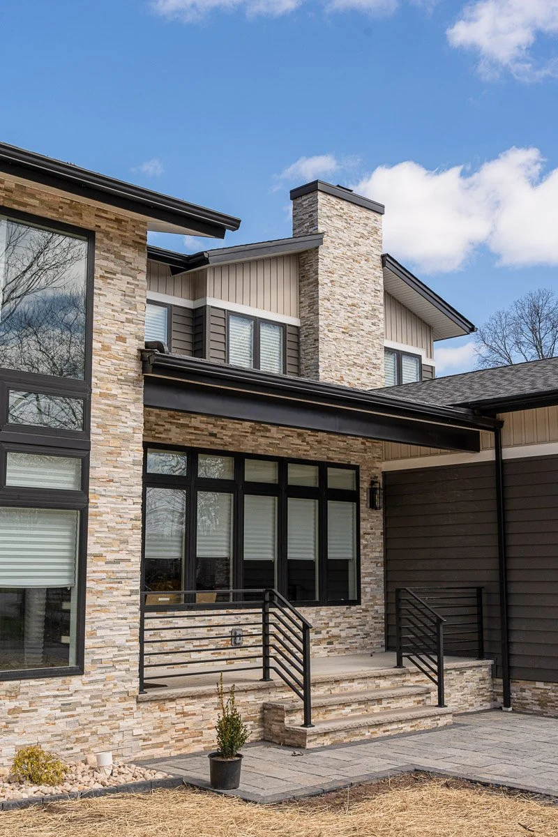 A modern house with stone and siding exterior, black window frames, a small front porch with steps, and a clear blue sky.