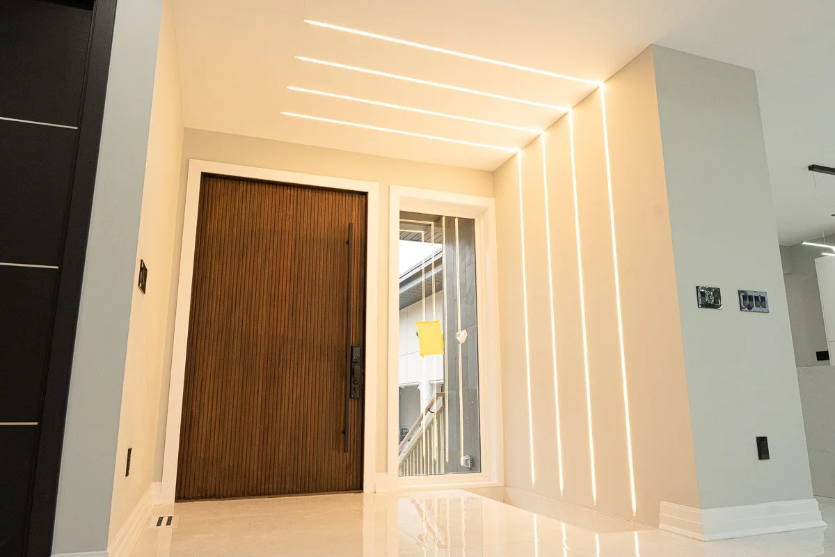 Modern entryway with a wooden front door and a glass side panel, illuminated by LED strip lights on the ceiling and wall, with a light-colored floor and walls.