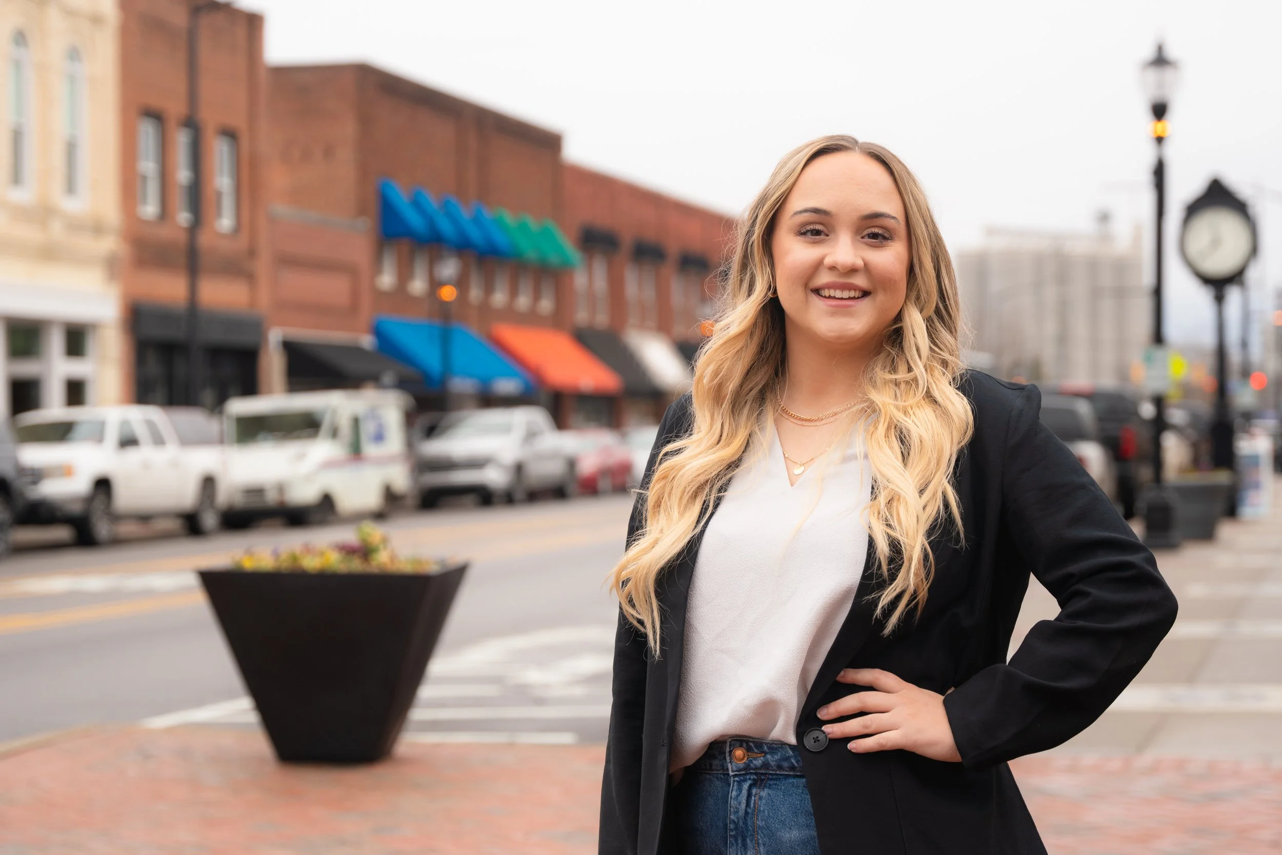 A young woman with long blonde hair smiling and posing outdoors in a city street, wearing a black blazer, white shirt, and blue jeans.