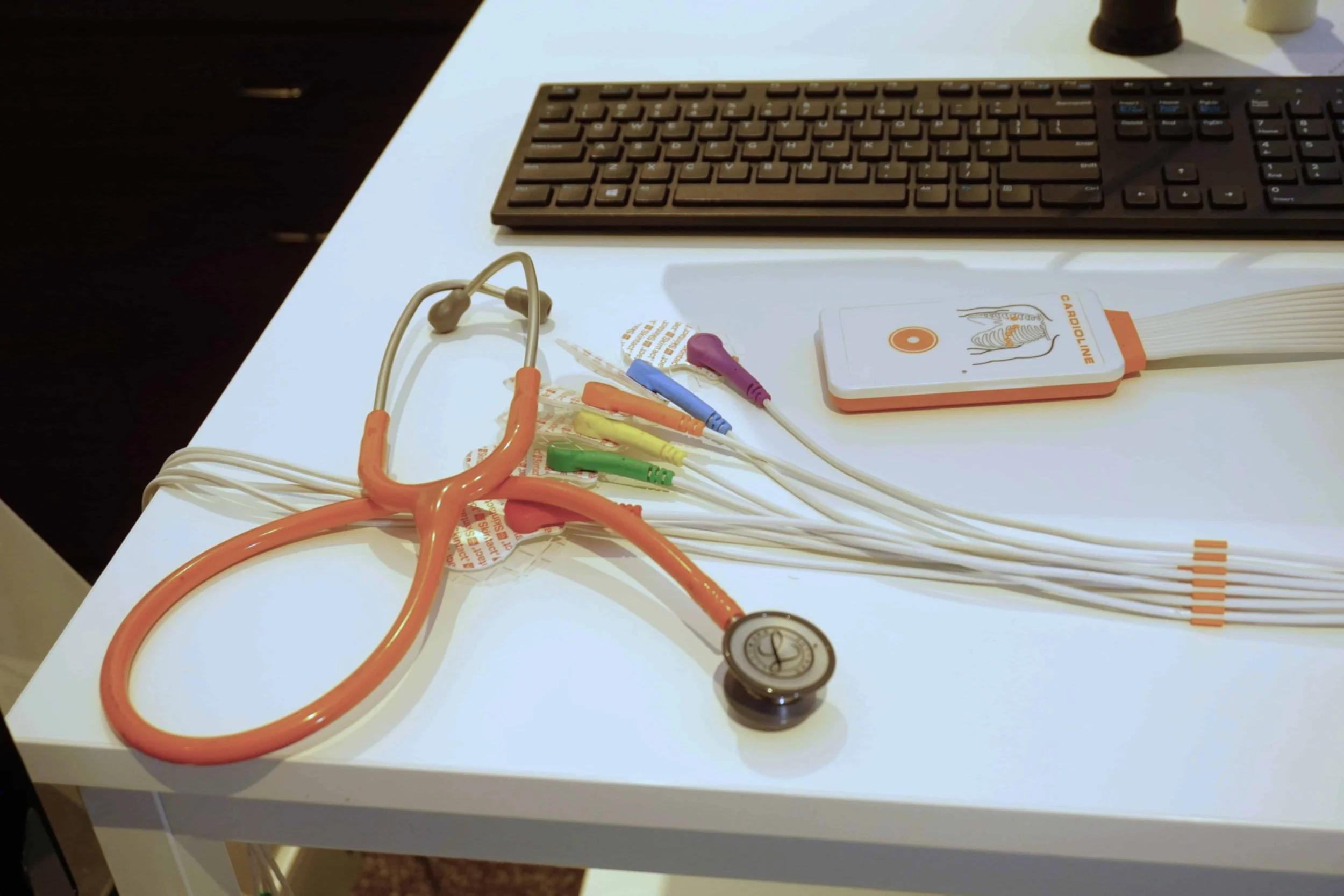 A white medical table with a stethoscope, a blood pressure cuff, orange, green, blue, yellow, and purple cables with connectors, a white book with orange accents, and a black keyboard on it.