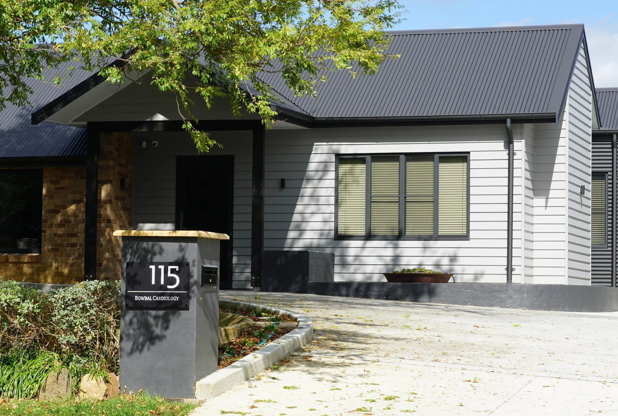 Modern house with white siding and black accents, front yard with greenery, large window with blinds, and a sign for Bowral Cardiology at the street corner.