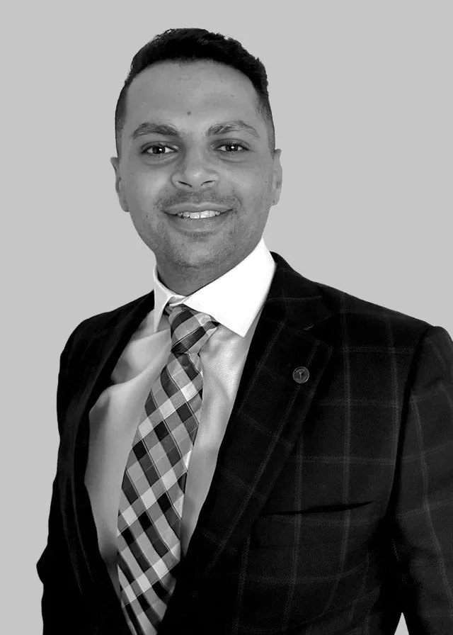 A man wearing a plaid tie, white shirt, and dark blazer, smiling, with a plain gray background.