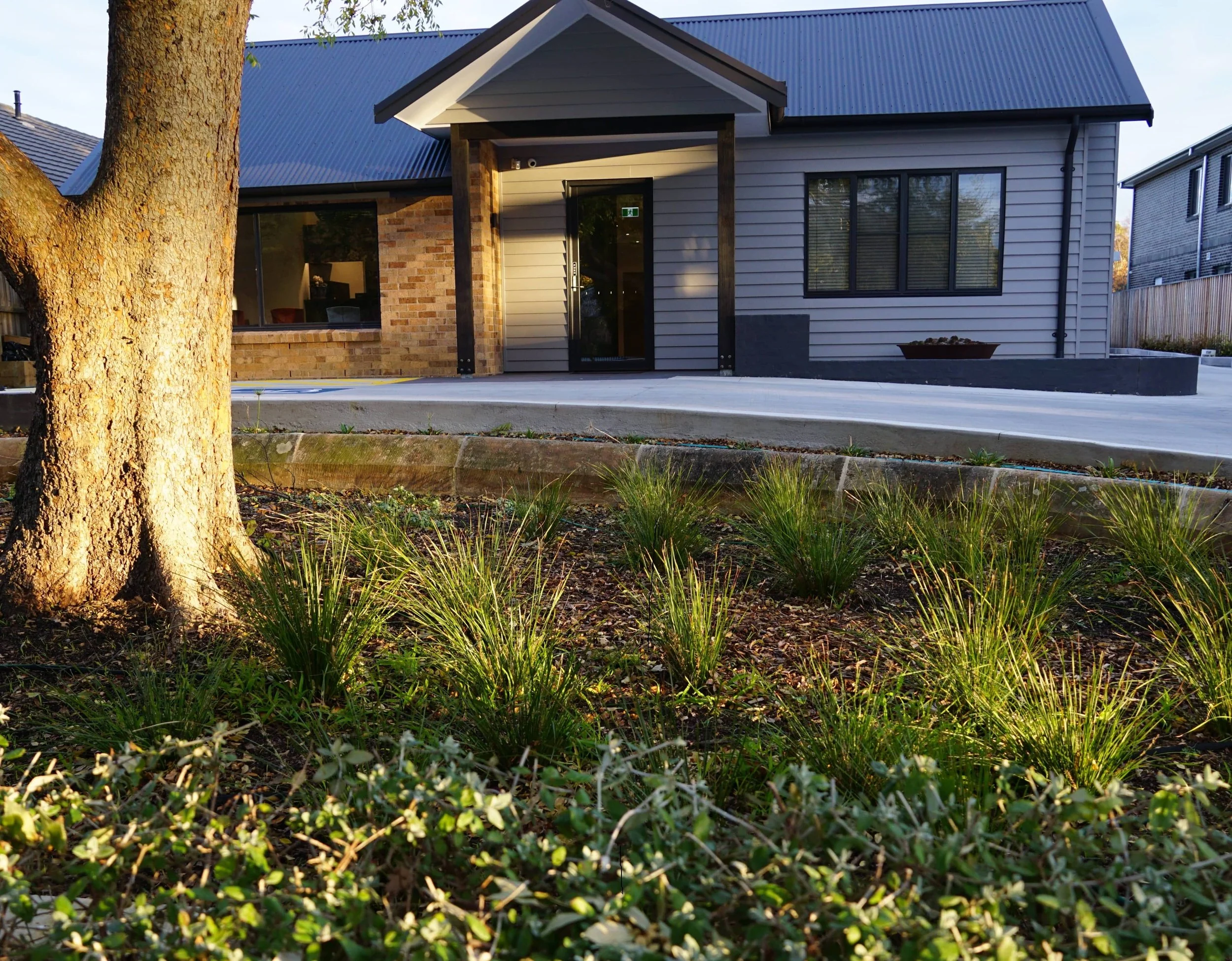 A residential house with a brick and siding exterior, large windows, a porch, and a blue metal roof, surrounded by greenery and trees.