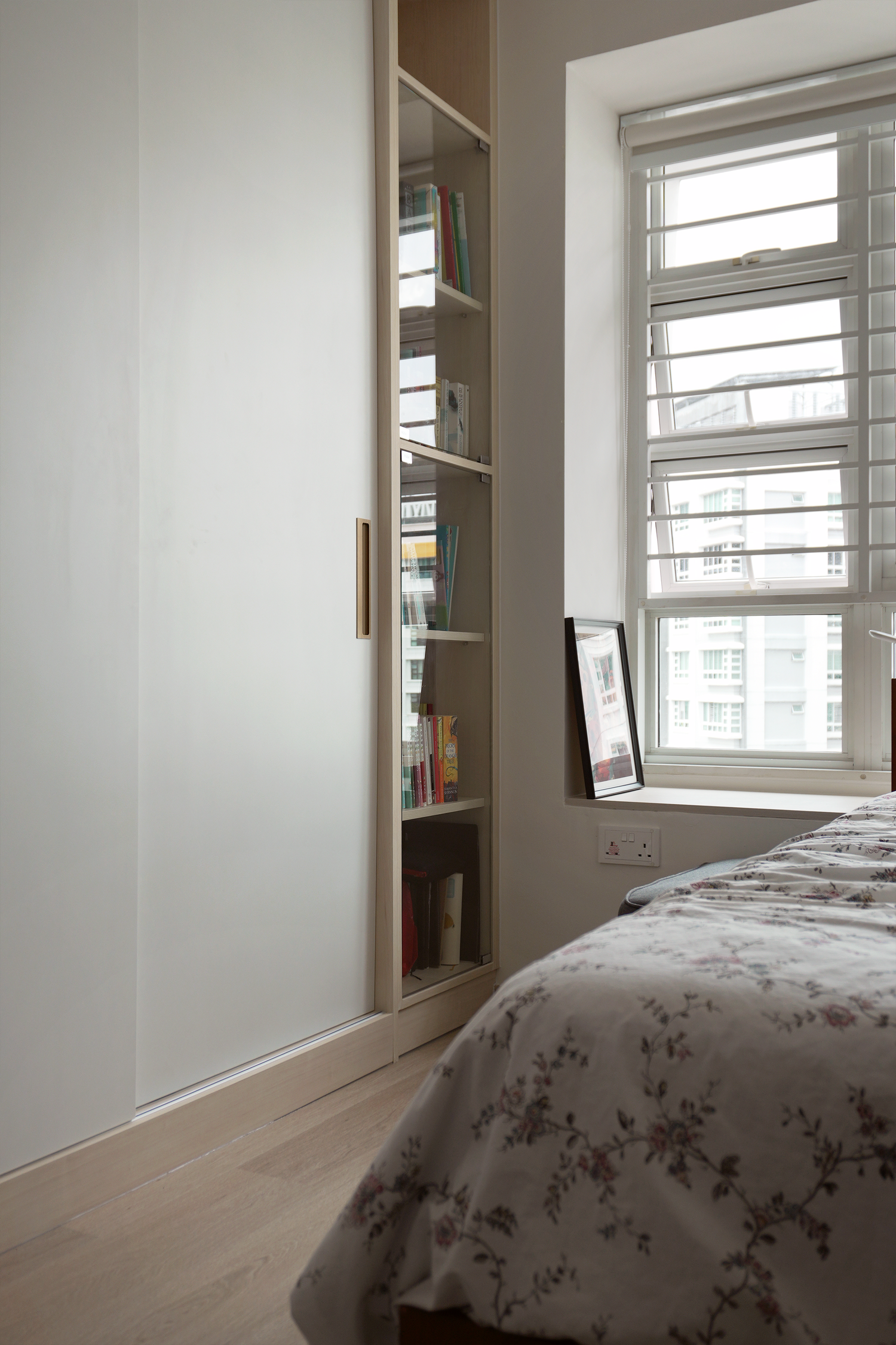 A bedroom with a white sliding door closet, a window with horizontal blinds, a framed picture resting on the windowsill, and a bed with a floral-patterned sheet.