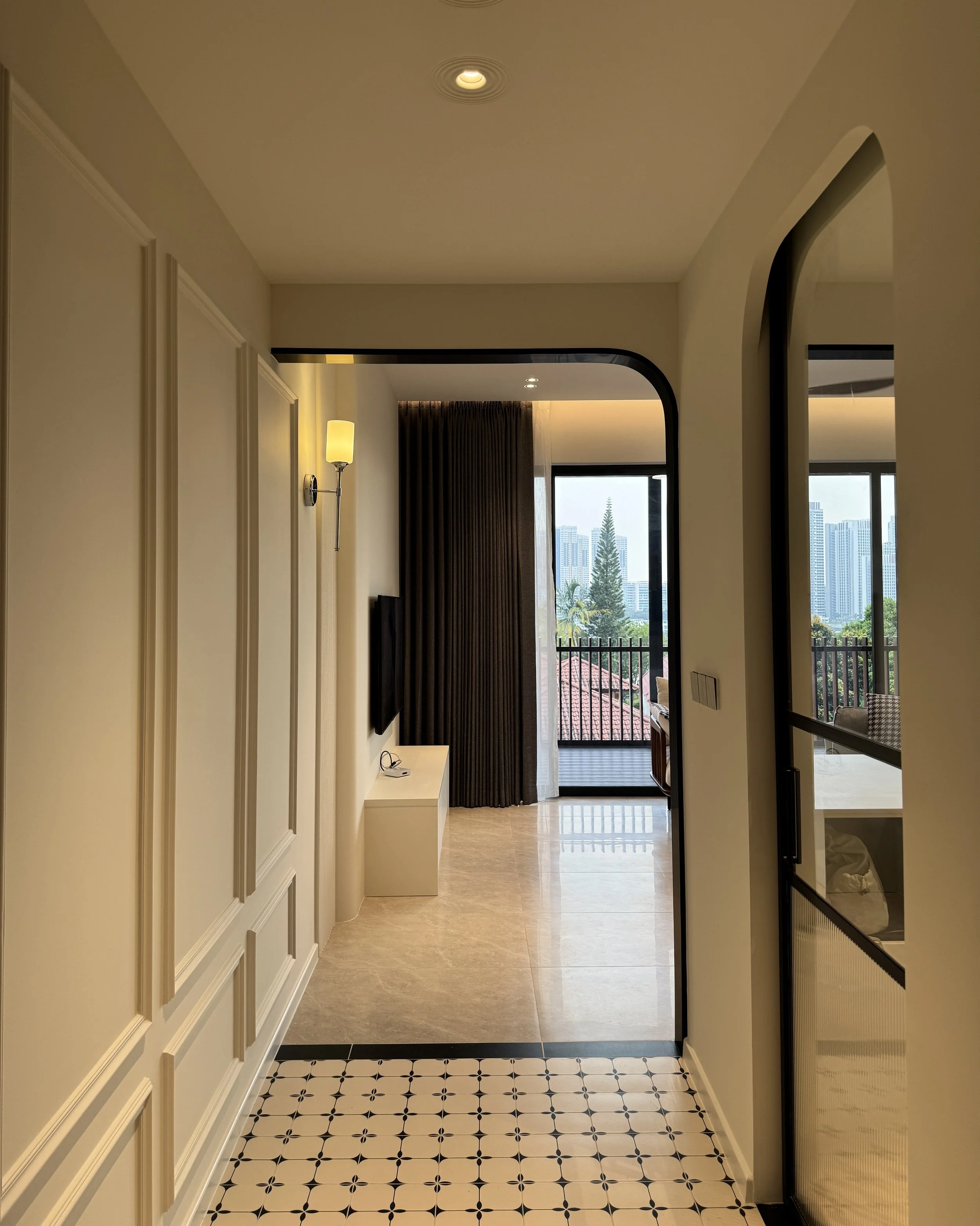 Interior view of a modern apartment corridor leading to a balcony with cityscape view, featuring beige walls, black and white patterned tile floor, and a wall-mounted lamp.