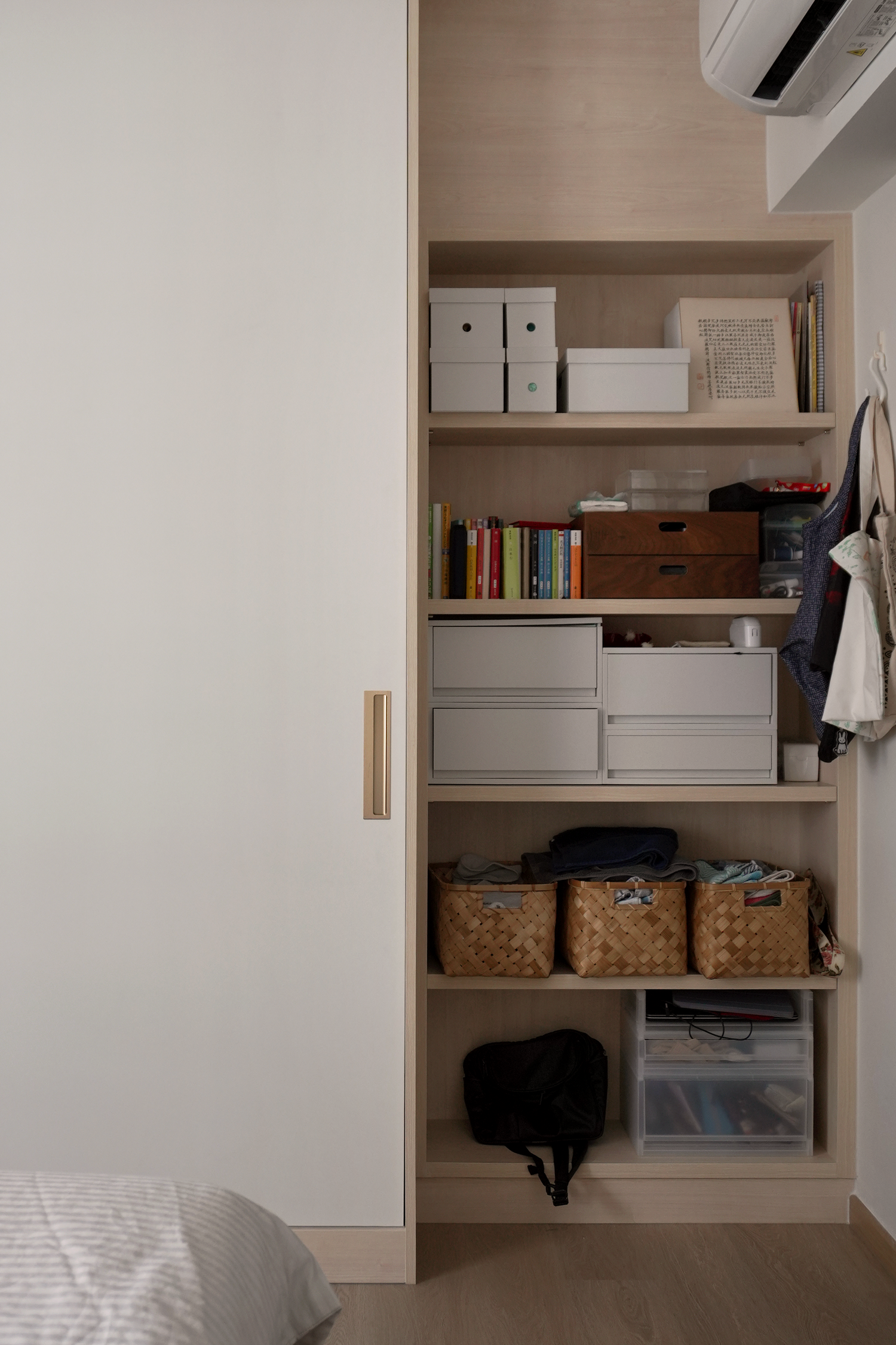A view of a bedroom with a built-in bookshelf and storage unit. The bookshelf contains books, boxes, and decorative items. Part of a bed with a striped blanket and pillow is visible in the foreground.