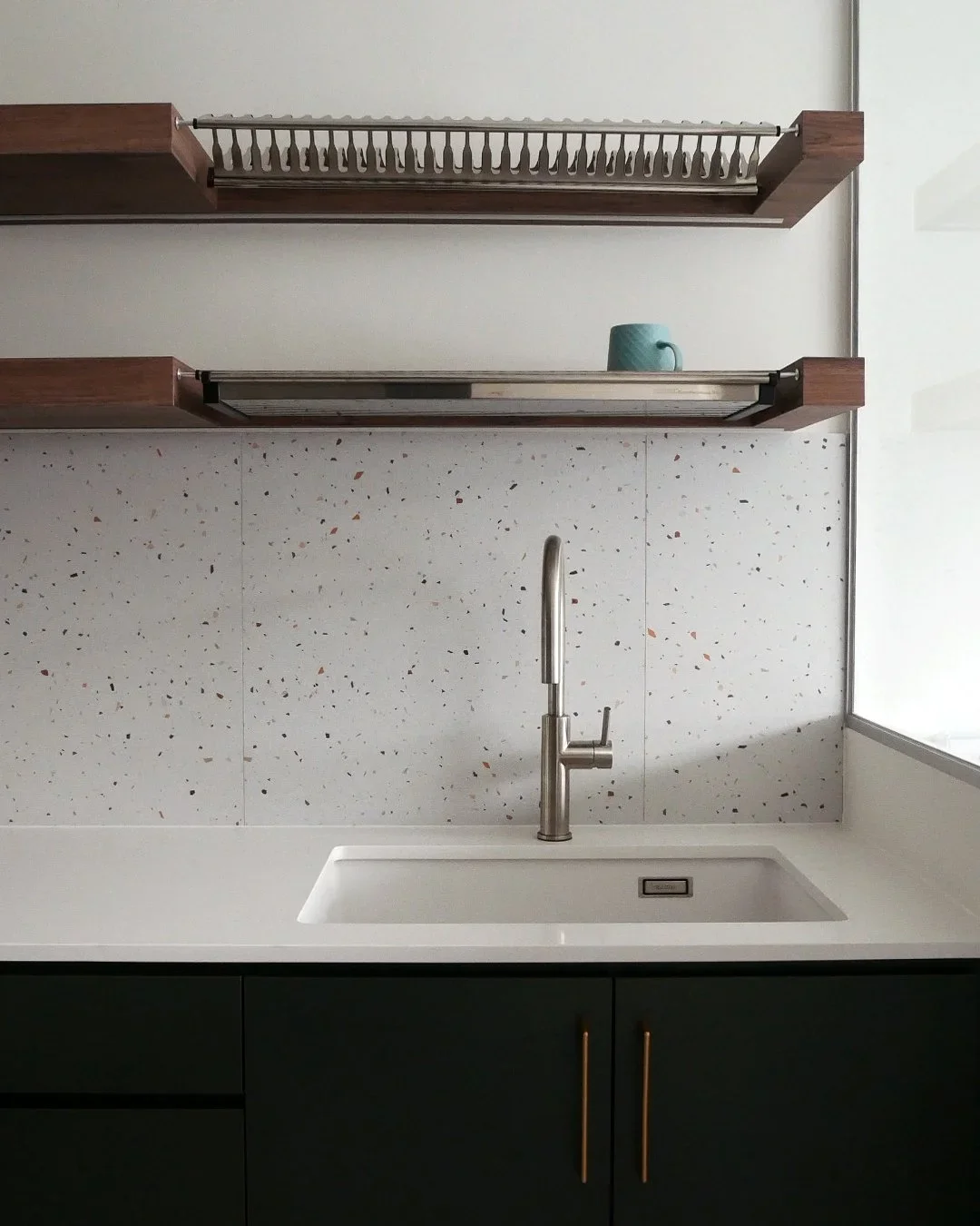 Modern kitchen sink with a metallic faucet, white countertop, and terrazzo backsplash, upper wooden shelves, and a small teal mug.