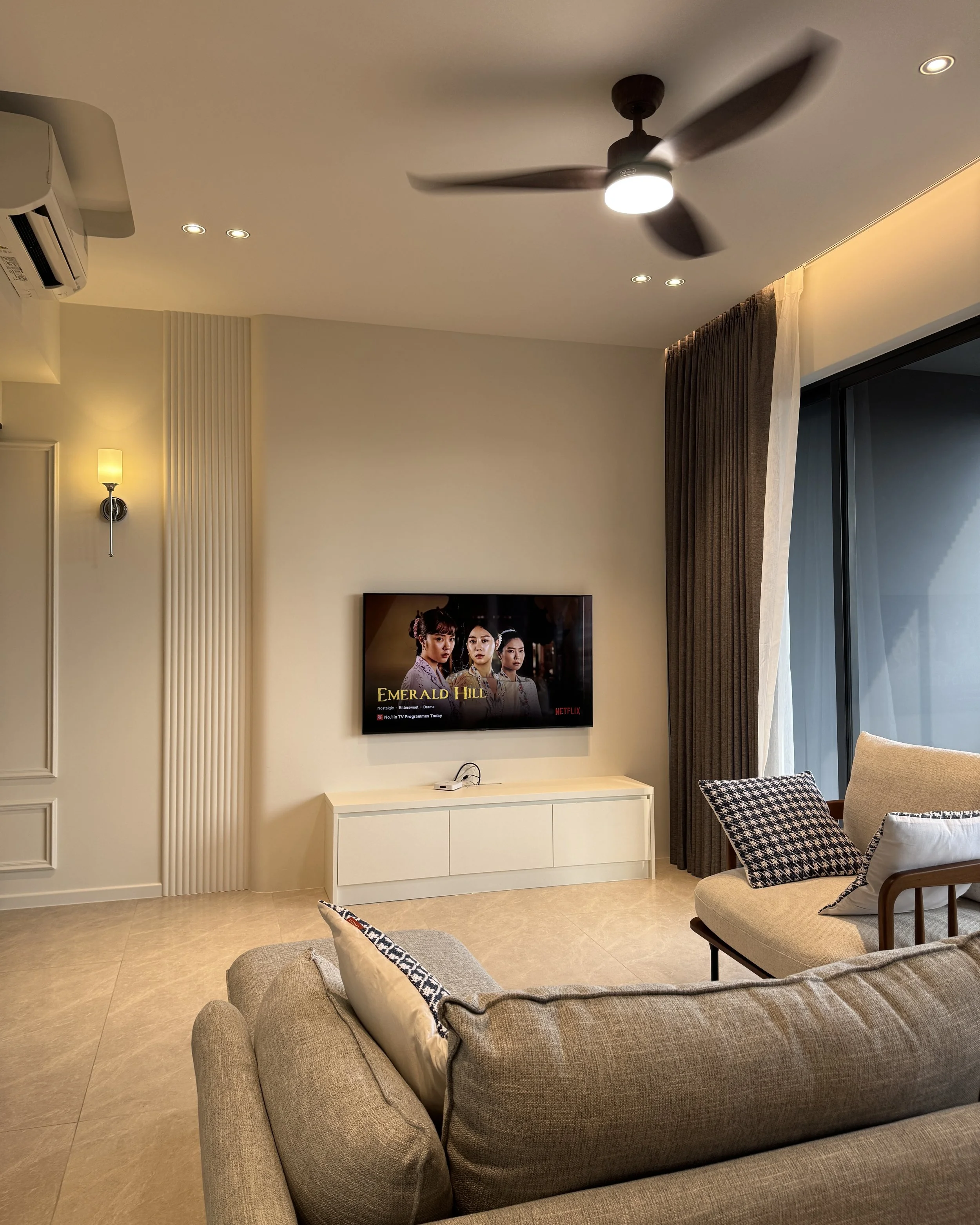 Living room with beige sofa, pillows, wall-mounted TV, cream and brown curtains, and ceiling fan with light.