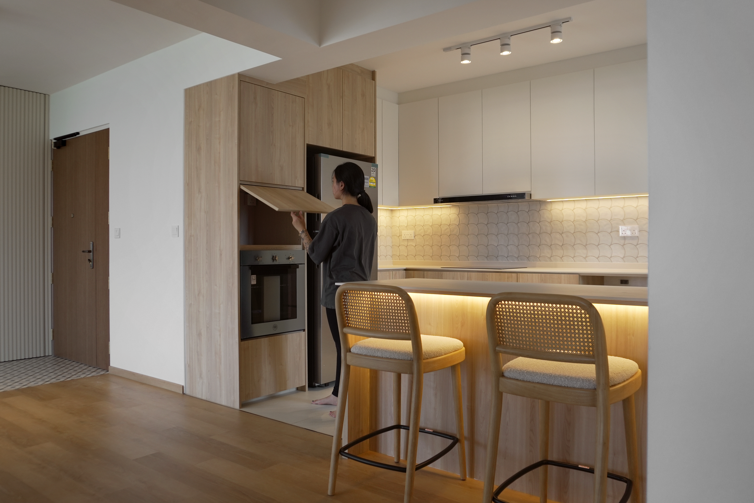 A woman in a black shirt and black leggings standing in a modern kitchen, opening a cabinet near a built-in oven. The kitchen has white upper cabinets, a scalloped tile backsplash, wooden lower cabinets, and a ceiling track light. The foreground show