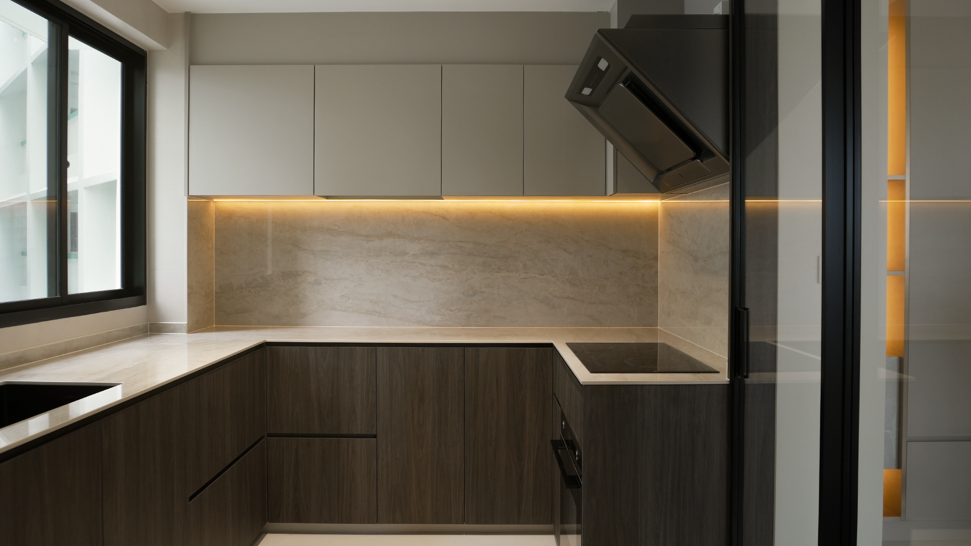 Modern kitchen with dark lower cabinets, light upper cabinets, and a beige marble backsplash illuminated by under-cabinet lighting. Includes a black induction cooktop, a black range hood, and a window showing the outdoor structure.
