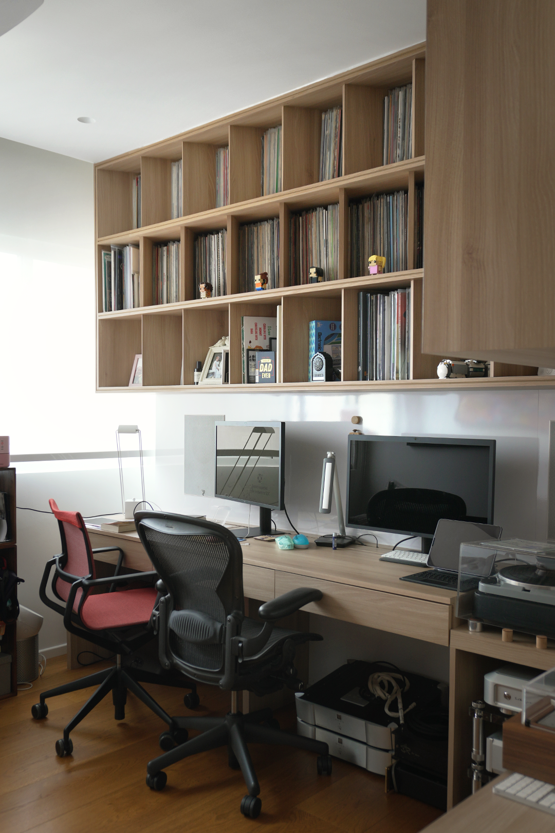 Home office with two desks, four chairs, dual monitors, a record player, a wooden bookshelf with vinyl records and small decorative items, and a white wall.