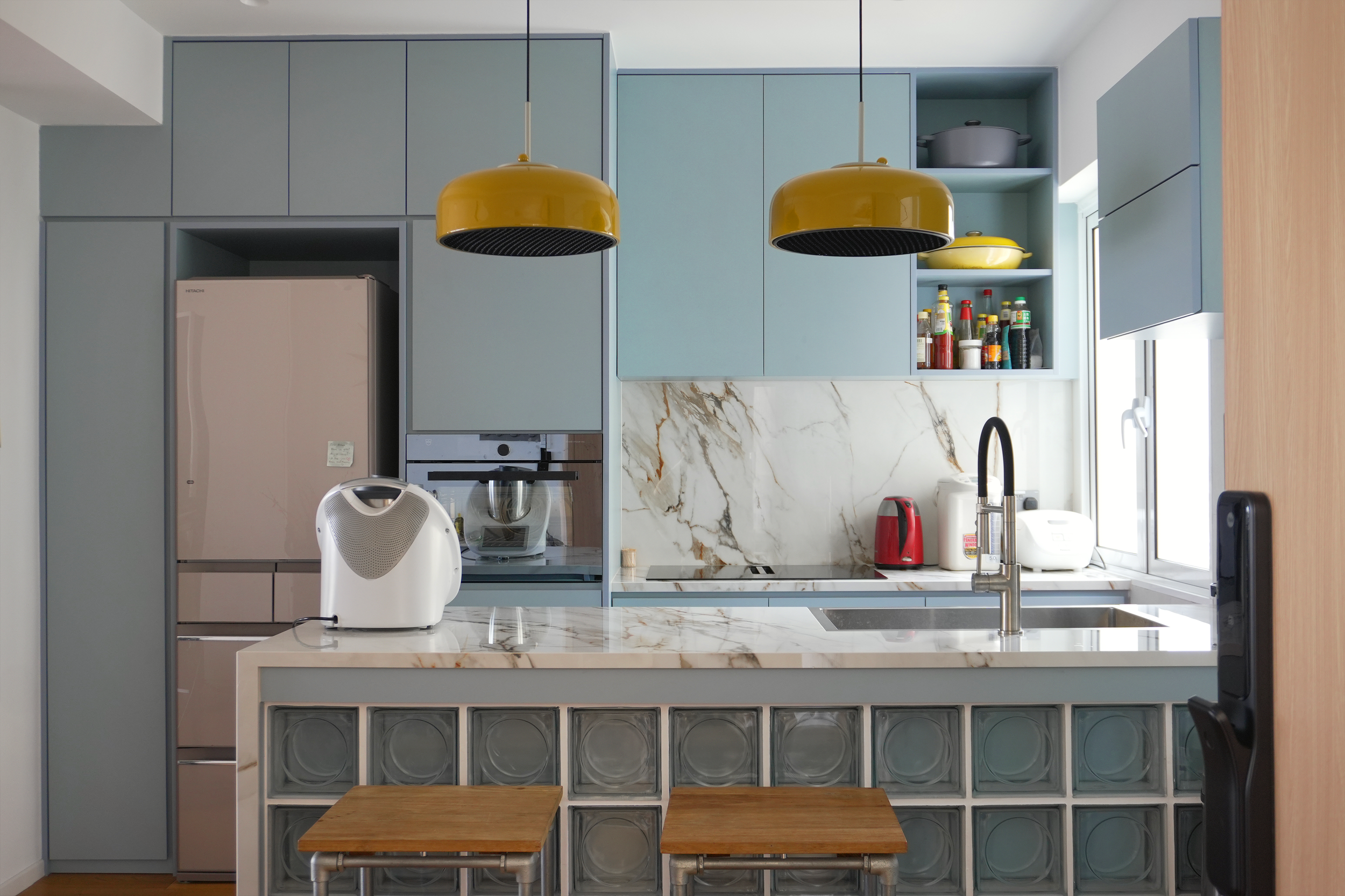 Modern kitchen with blue cabinets, marble countertop, yellow pendant lights, appliances, and open shelving with bottles and containers