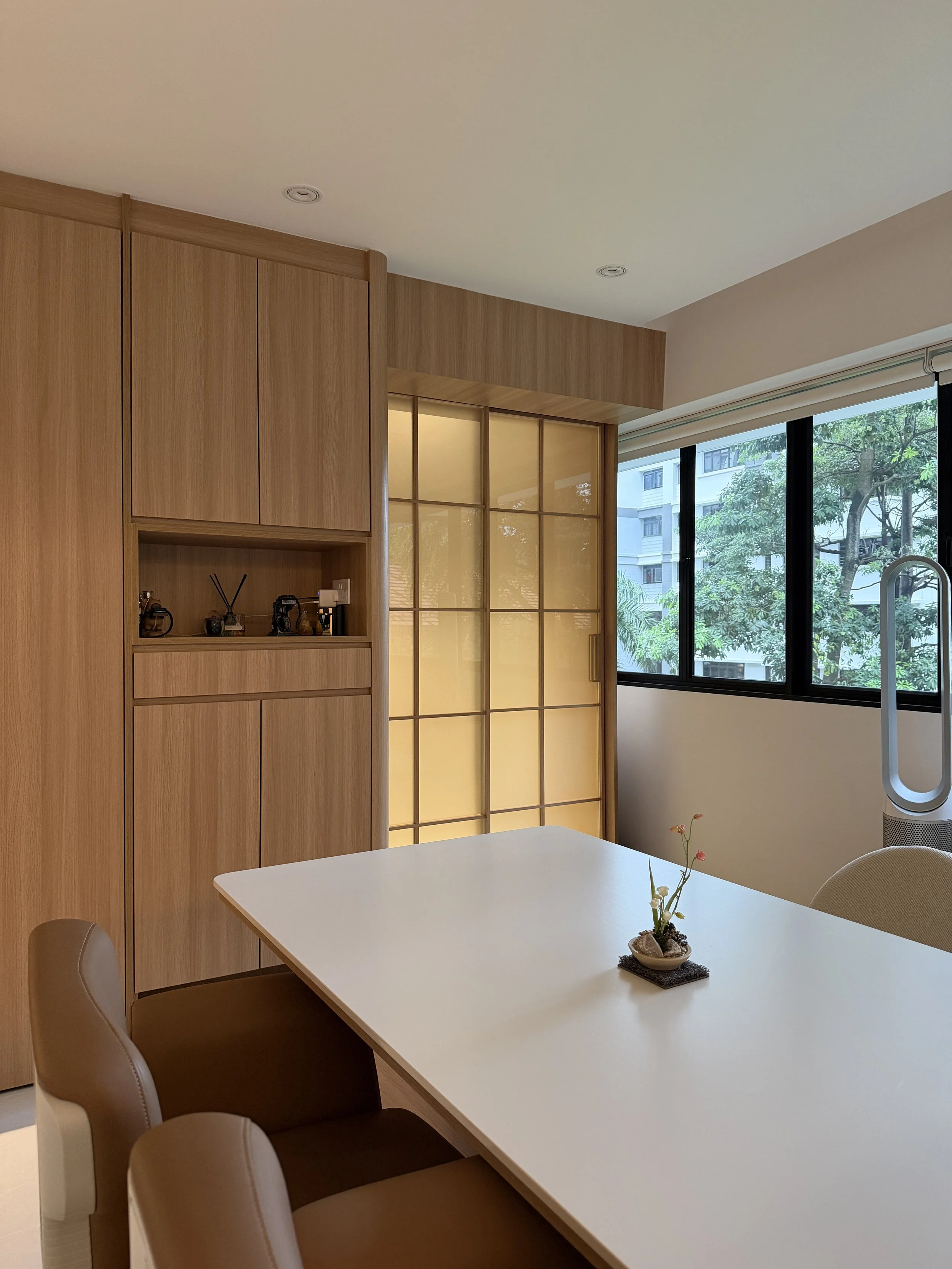 Modern dining room with a white table, brown chairs, wooden cabinets, a decorative plant on the table, large windows with a view of trees and buildings outside, and a Dyson fan near the window.