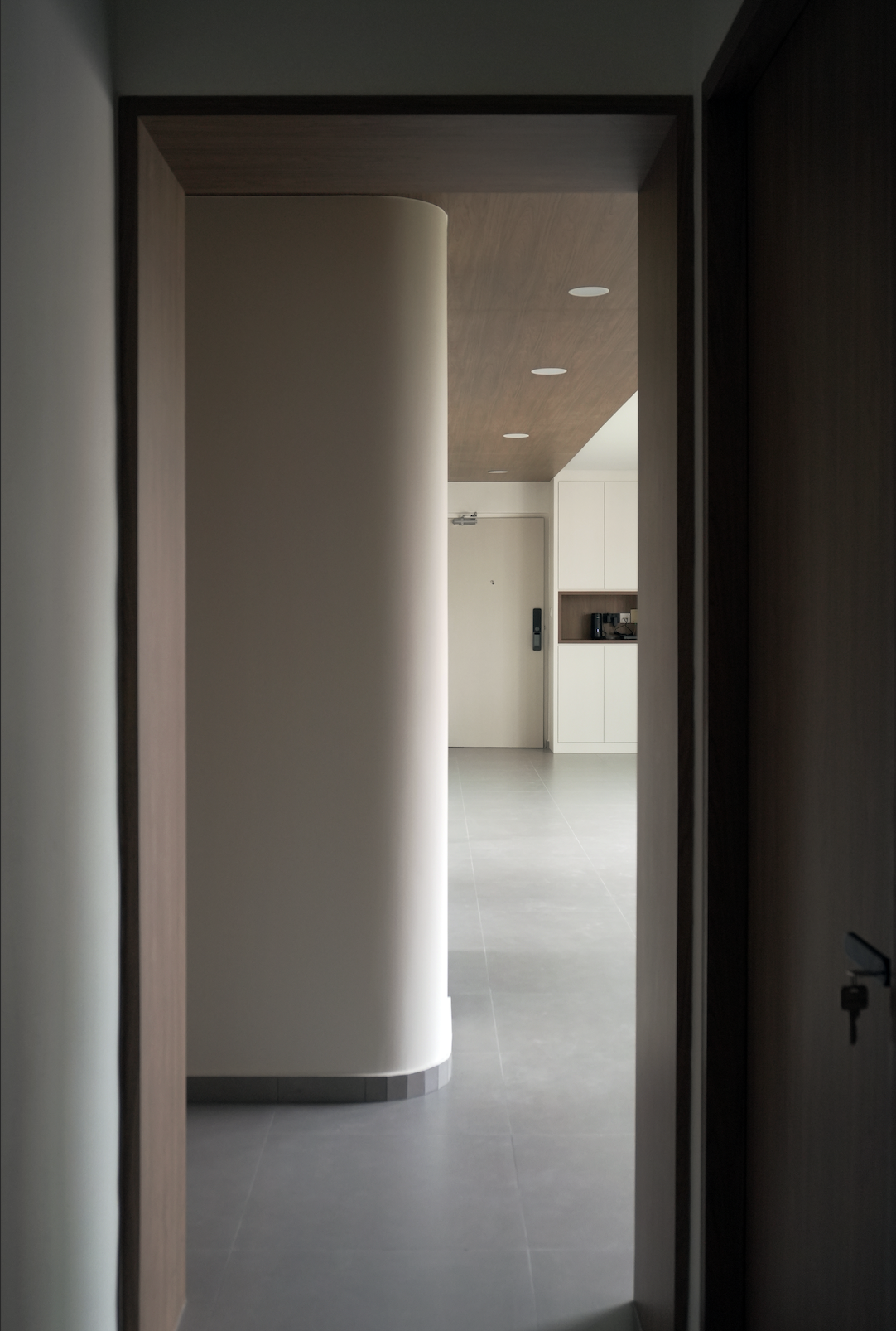 View of an interior space through a doorway, featuring a curved white wall, a wooden ceiling with recessed lighting, gray tiled floor, and a white door with a keypad lock in the background.