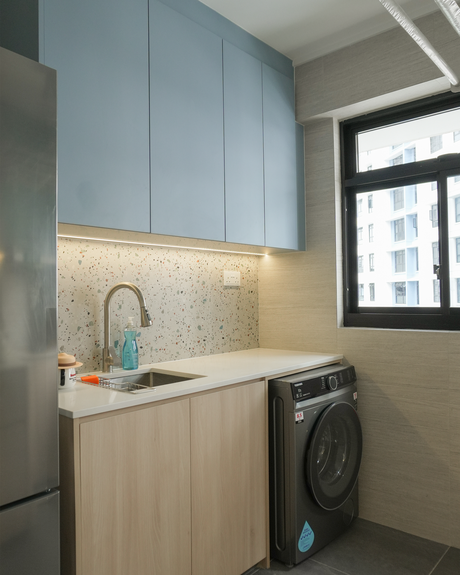 A modern laundry area with light wood cabinetry, a terrazzo backsplash, a white countertop with a sink, a soap dispenser, and an under-counter washing machine next to a window with a black frame, highlighting city buildings outside.