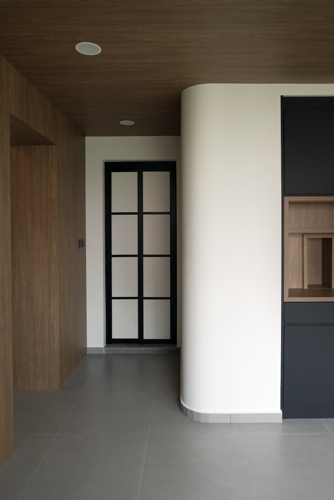 A modern interior hallway with wood-paneled ceiling and wall, a black framed glass door, and a curved white wall with built-in shelves.