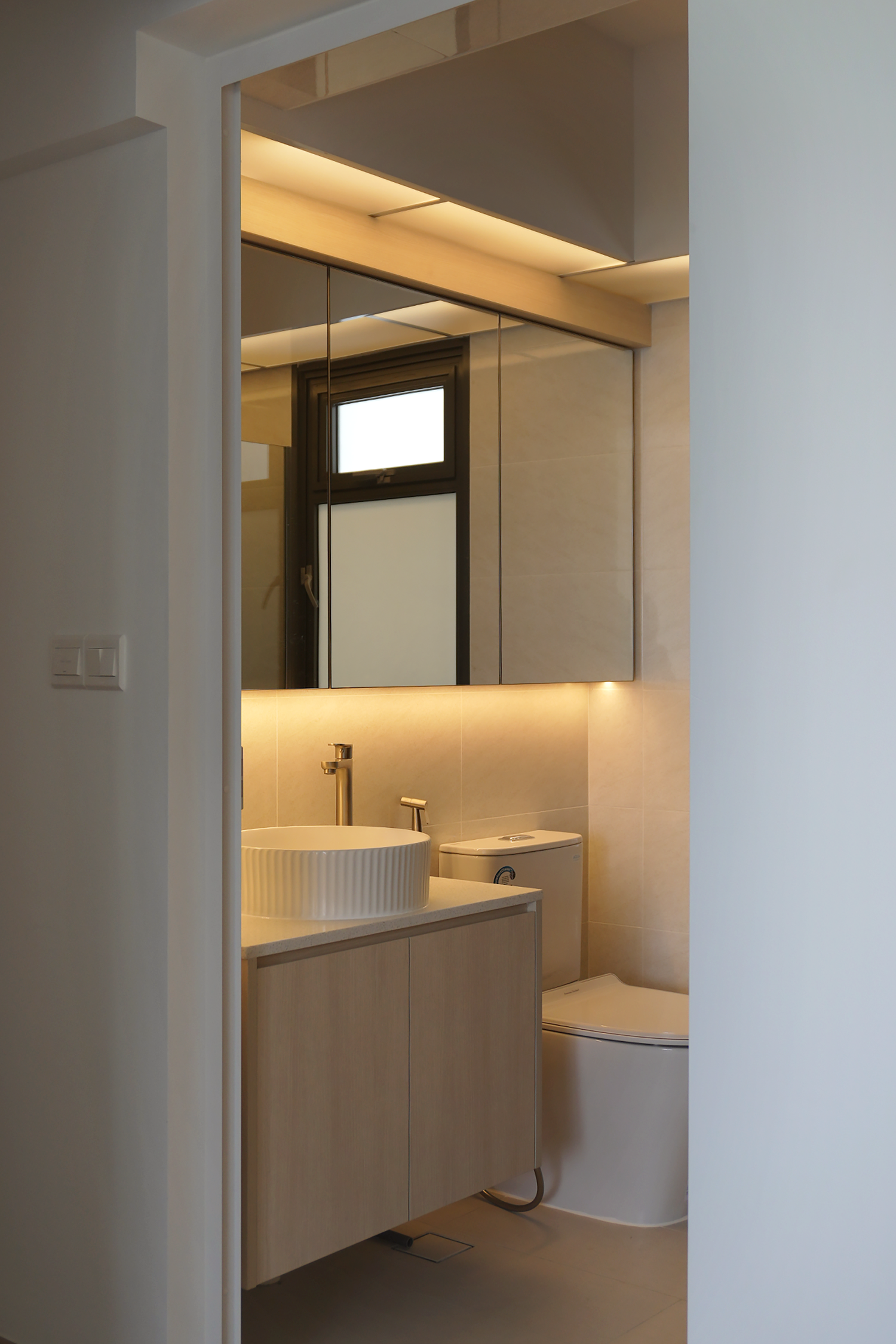 Modern bathroom with beige tiles, a small window, a vessel sink with a gold faucet, a wooden vanity, a toilet, and a shower enclosure.