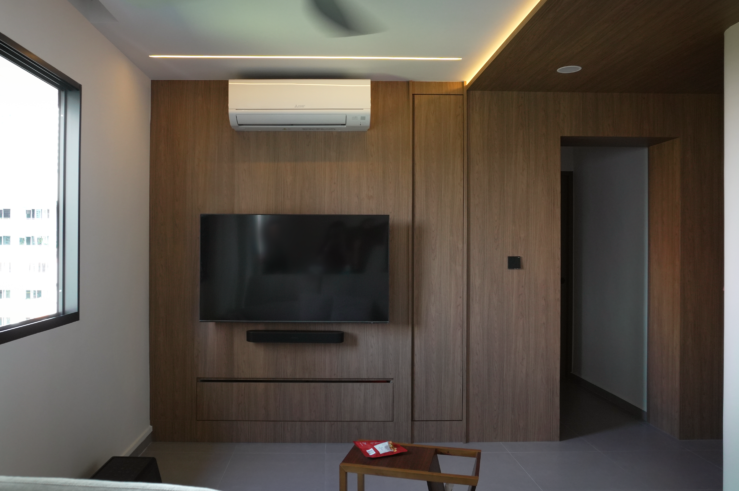 A modern living room with wooden wall paneling, a wall-mounted flat-screen TV, a black soundbar, an air conditioner unit above the TV, a window on the left side, and a small wooden table with a snack on it in the foreground.