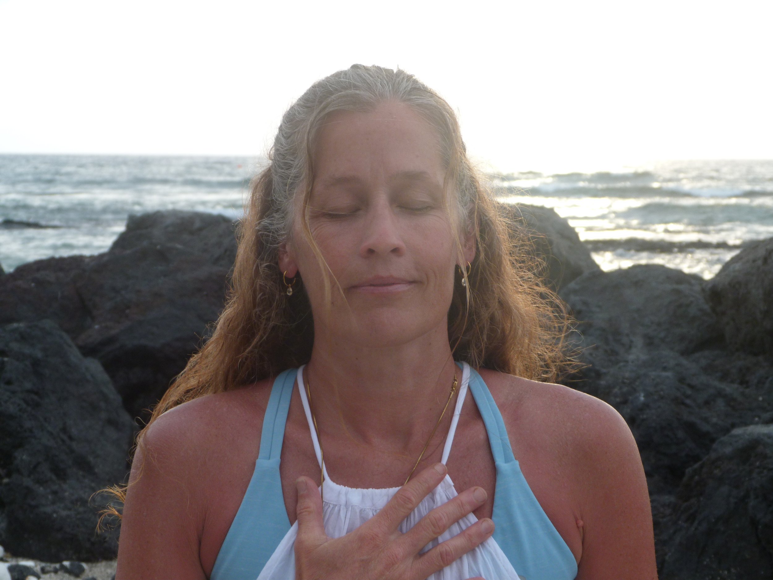 A woman with light brown curly hair, wearing a blue tank top and a white shirt, stands outdoors near the beach with her eyes closed and hand over her chest.