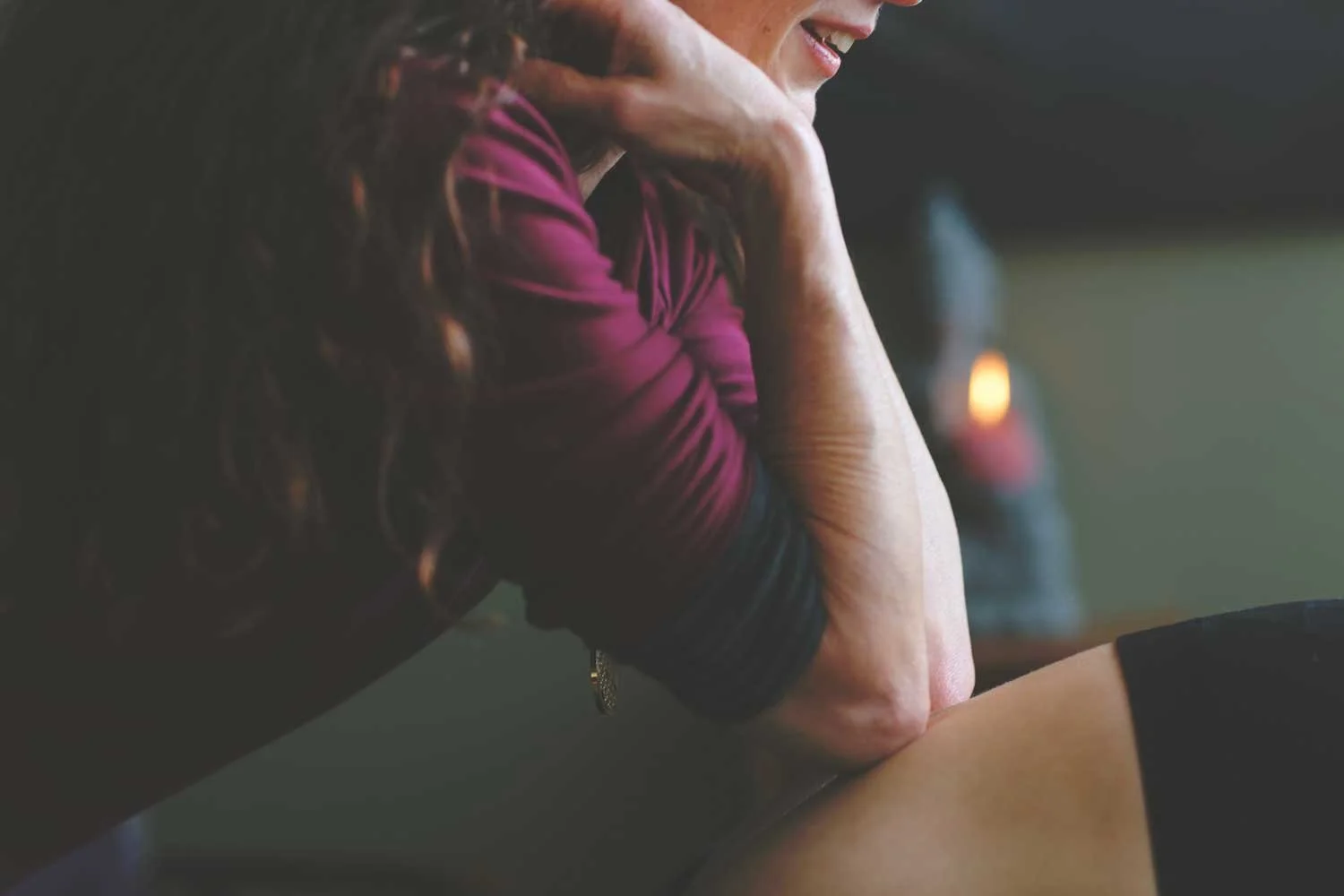 Close-up of a woman with curly hair resting her chin on her hand, smiling, in a dimly lit room.